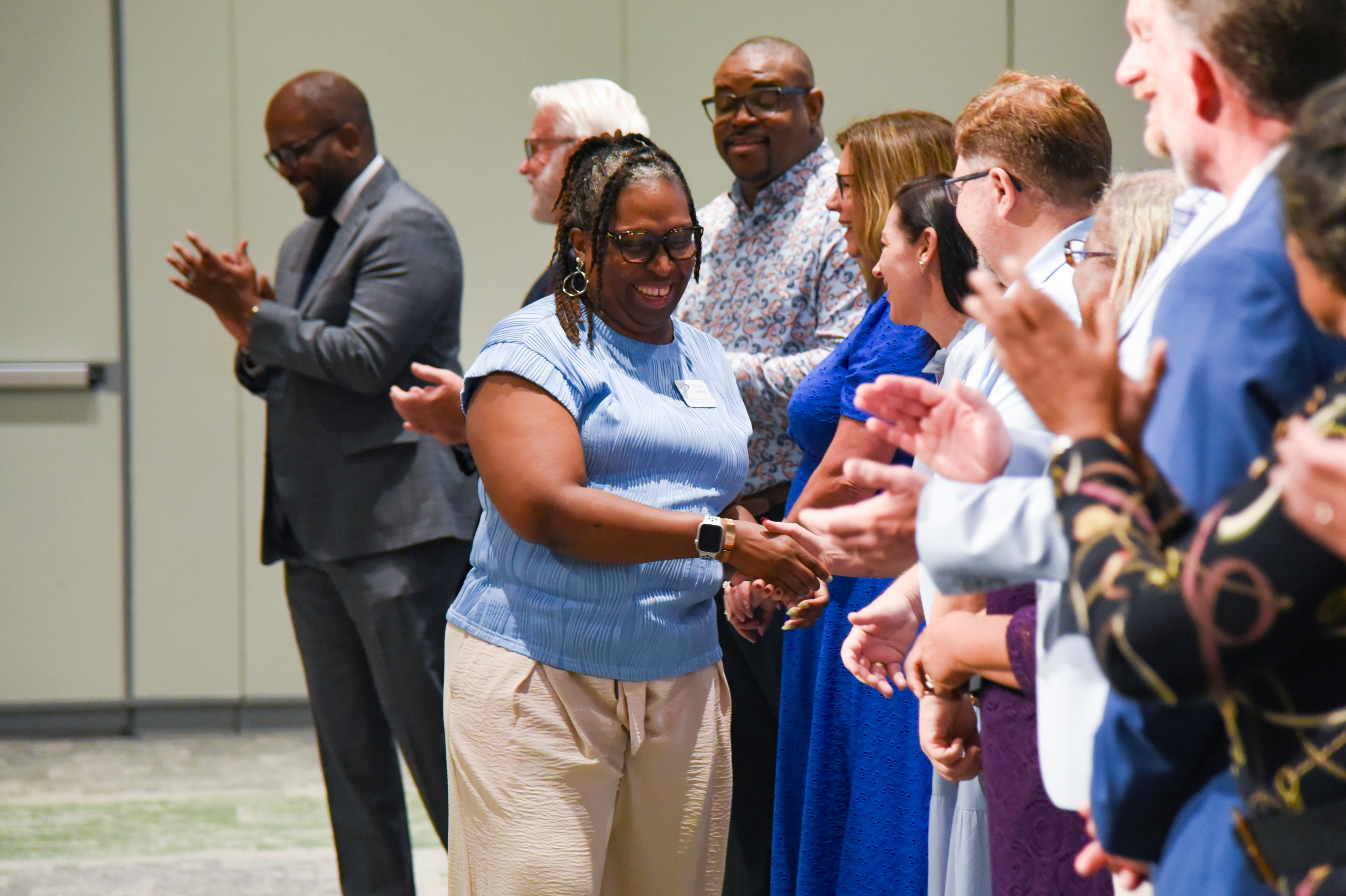 A woman in a light blue shirt and khaki pants stands among others clapping. Background shows a white wall.