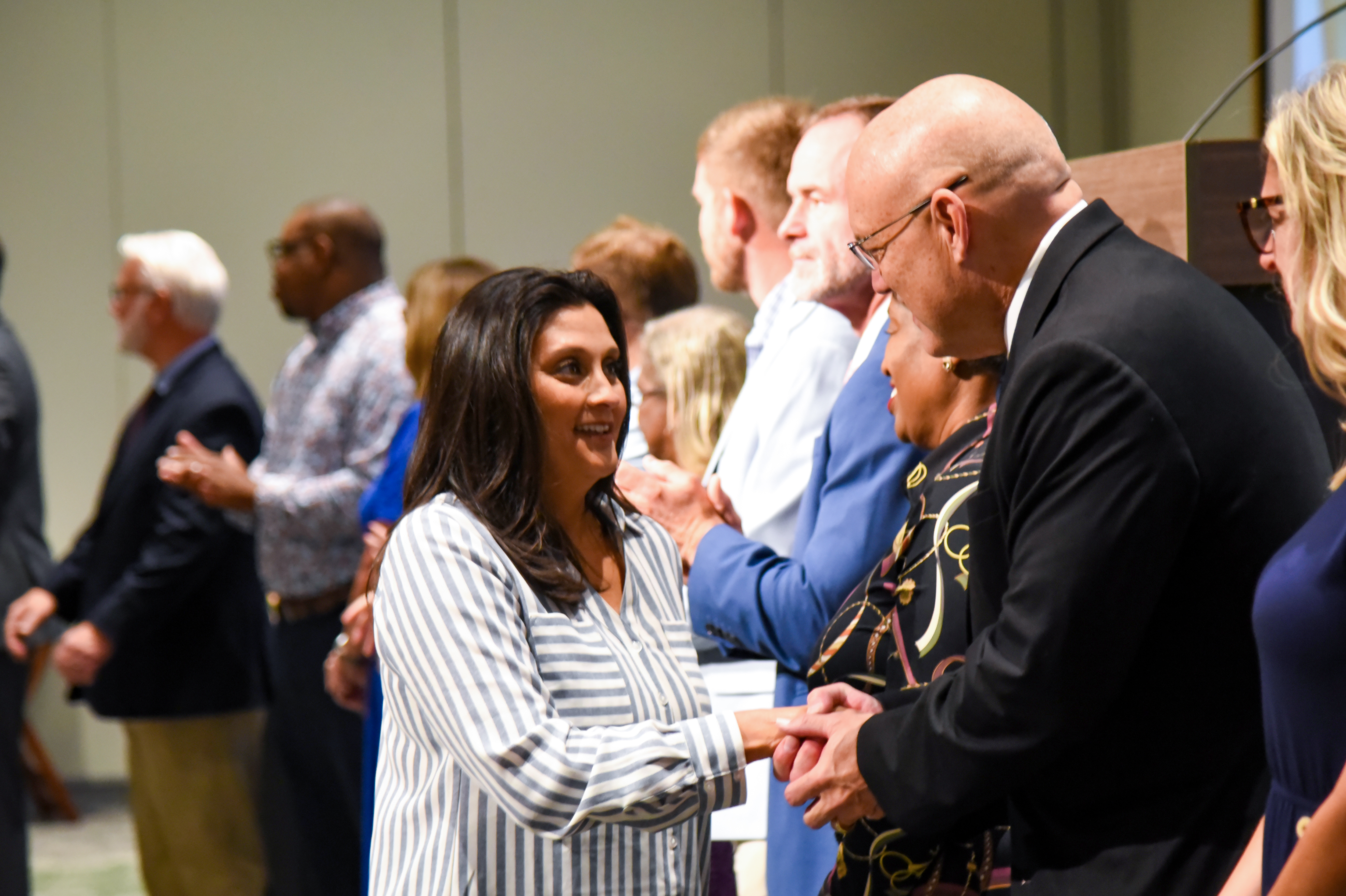 A woman in a striped shirt shakes hands with a man in a black suit at an indoor event.