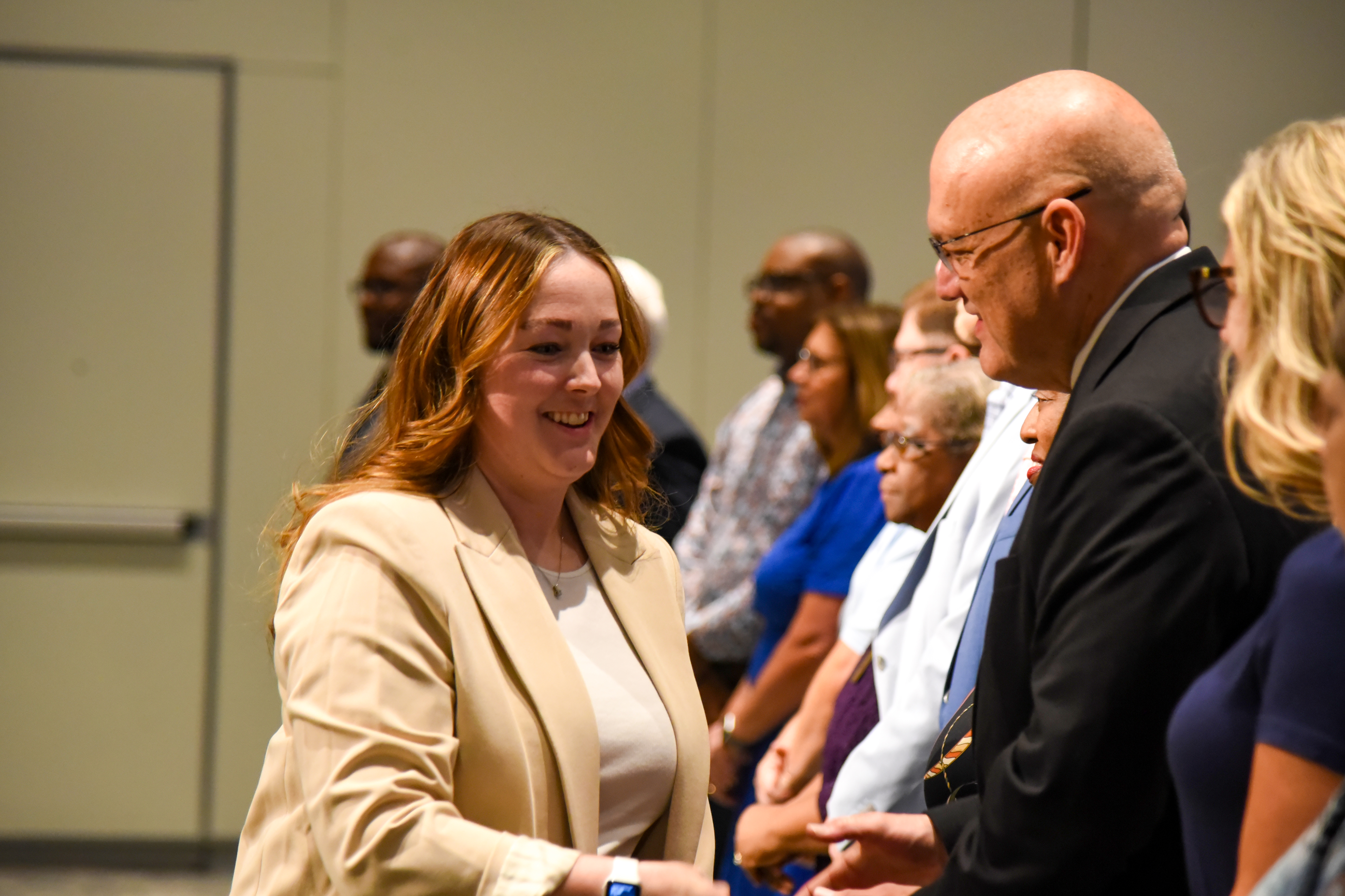 A group of people stand in a line. A woman in a white blouse and tan blazer smiles at a man.