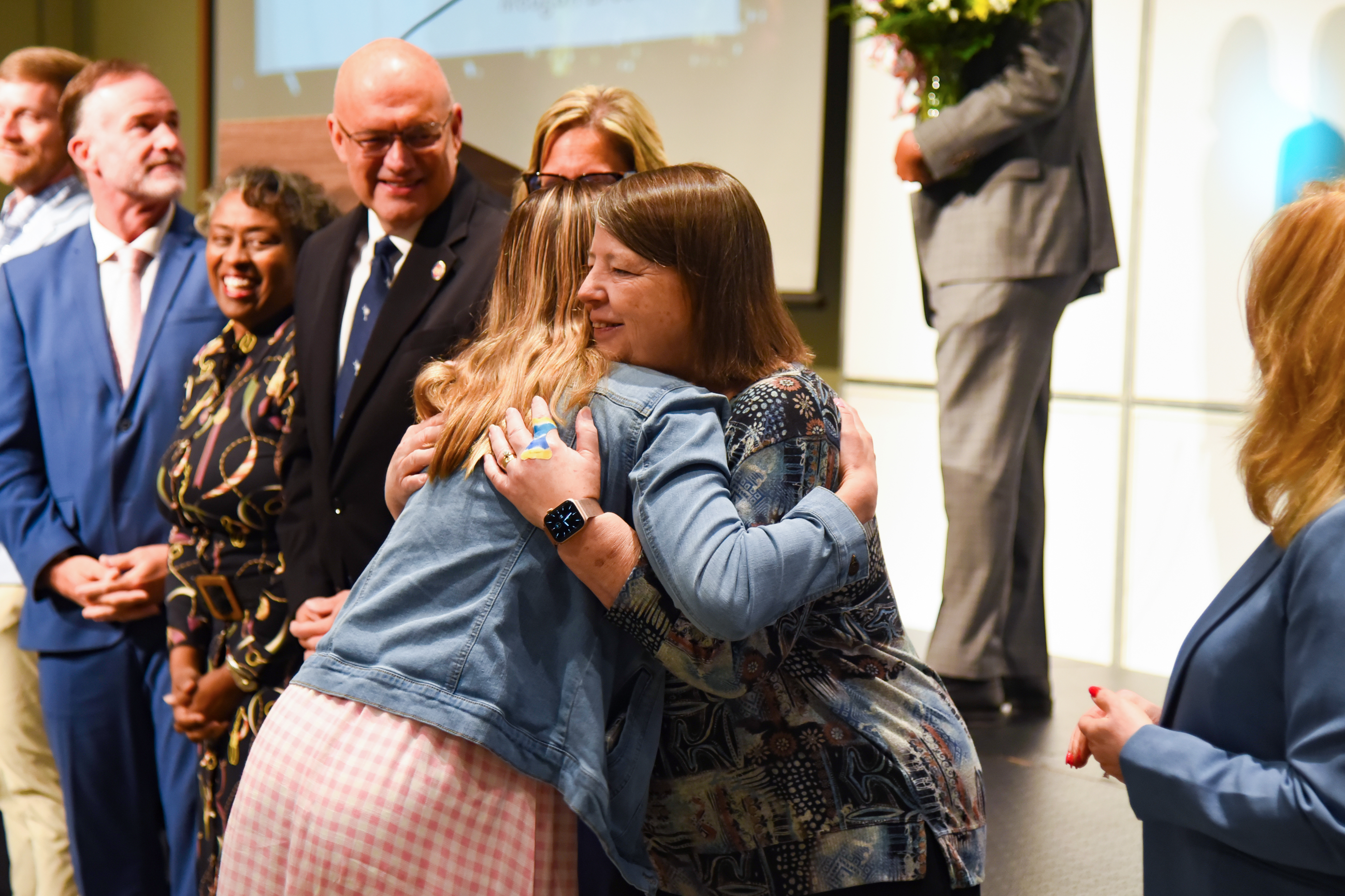 People gathered in a room. Two women hug each other; one has a floral top, and the other wears a denim jacket.