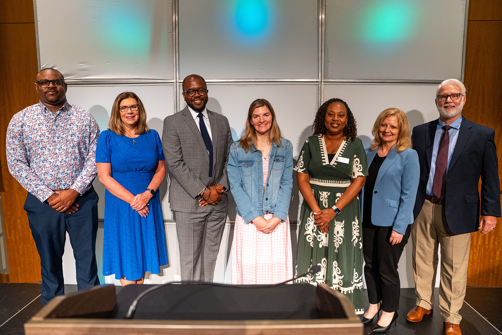 Seven adults stand in a row in a conference room. They are wearing business attire. The woman in the middle is smiling.