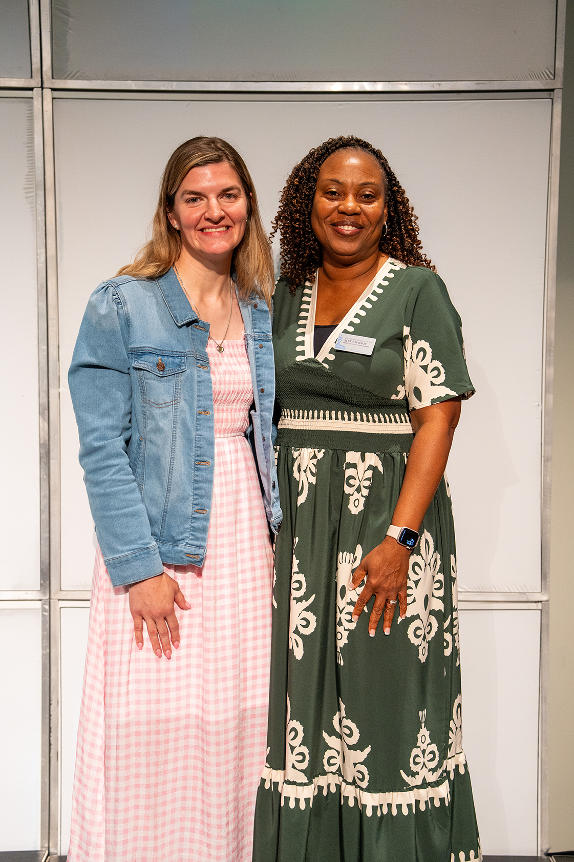 Two women stand smiling next to each other. One wears a denim jacket and pink dress, the other wears a green dress with white patterns.