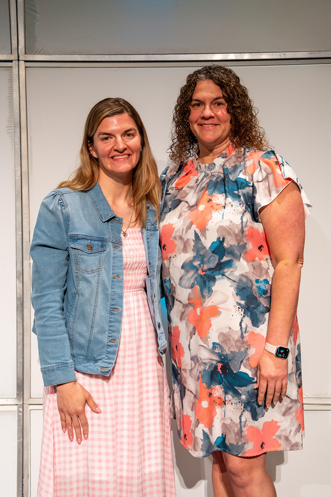 Two women stand next to each other, one in a pink dress and denim jacket, the other in a floral dress.