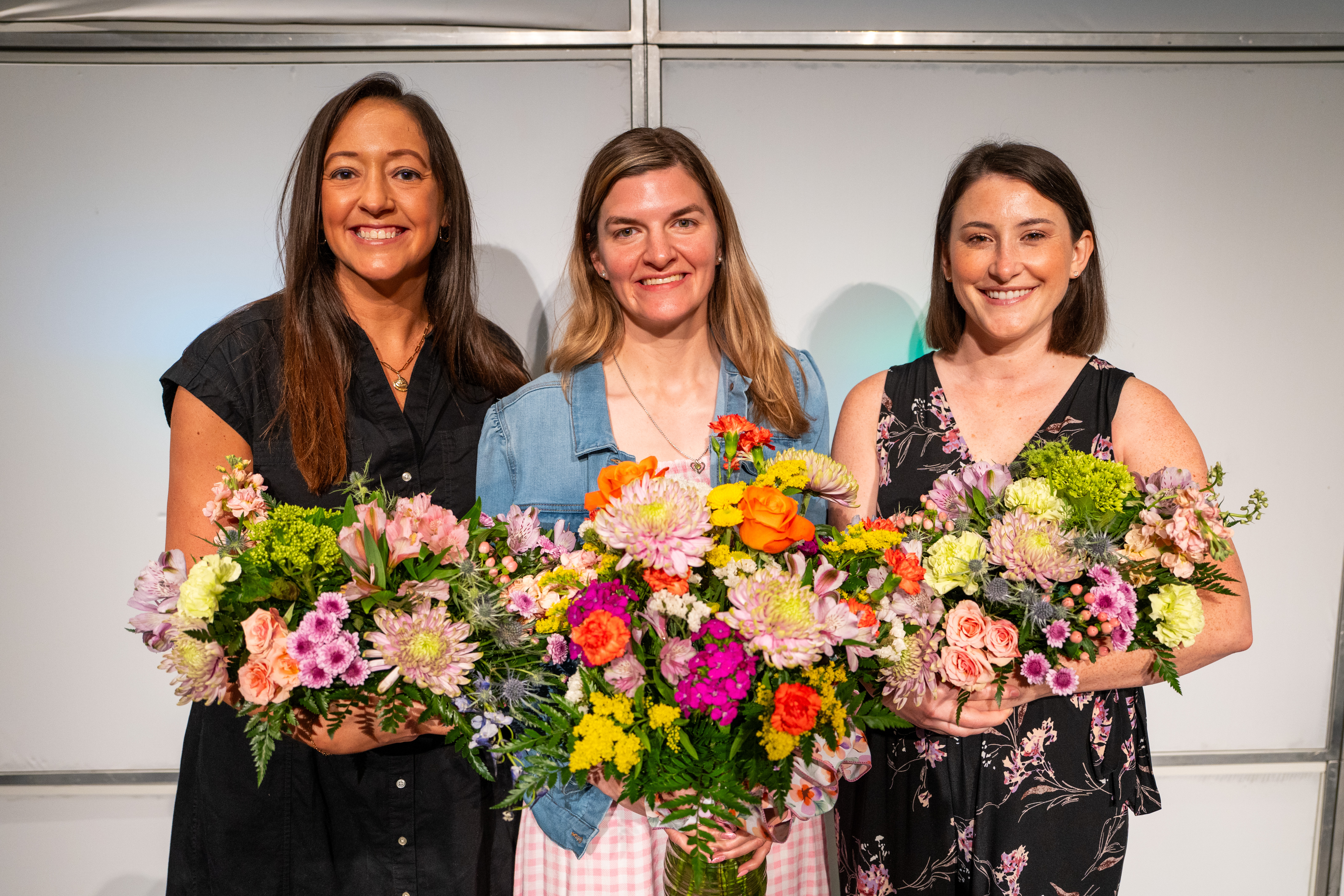 Three women hold colorful flower bouquets, smiling. One wears a black top, another a denim jacket, and the third a floral dress.
