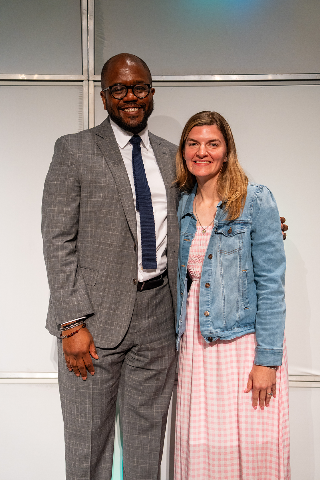 A man in a gray suit and a woman in a pink dress stand together, both smiling, against a white background.