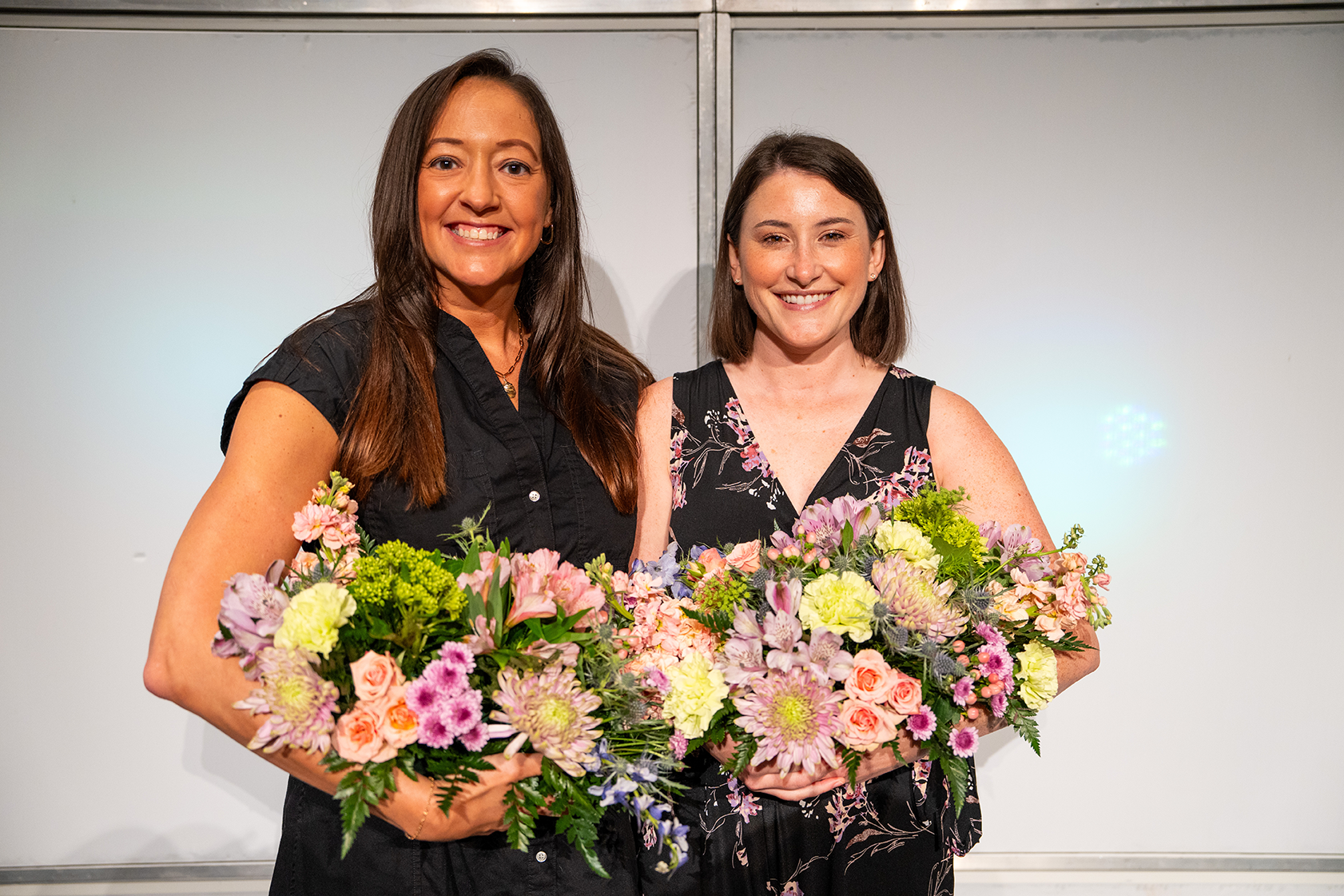 Two women in black dresses hold large floral bouquets. One woman smiles, and the other looks at the camera.