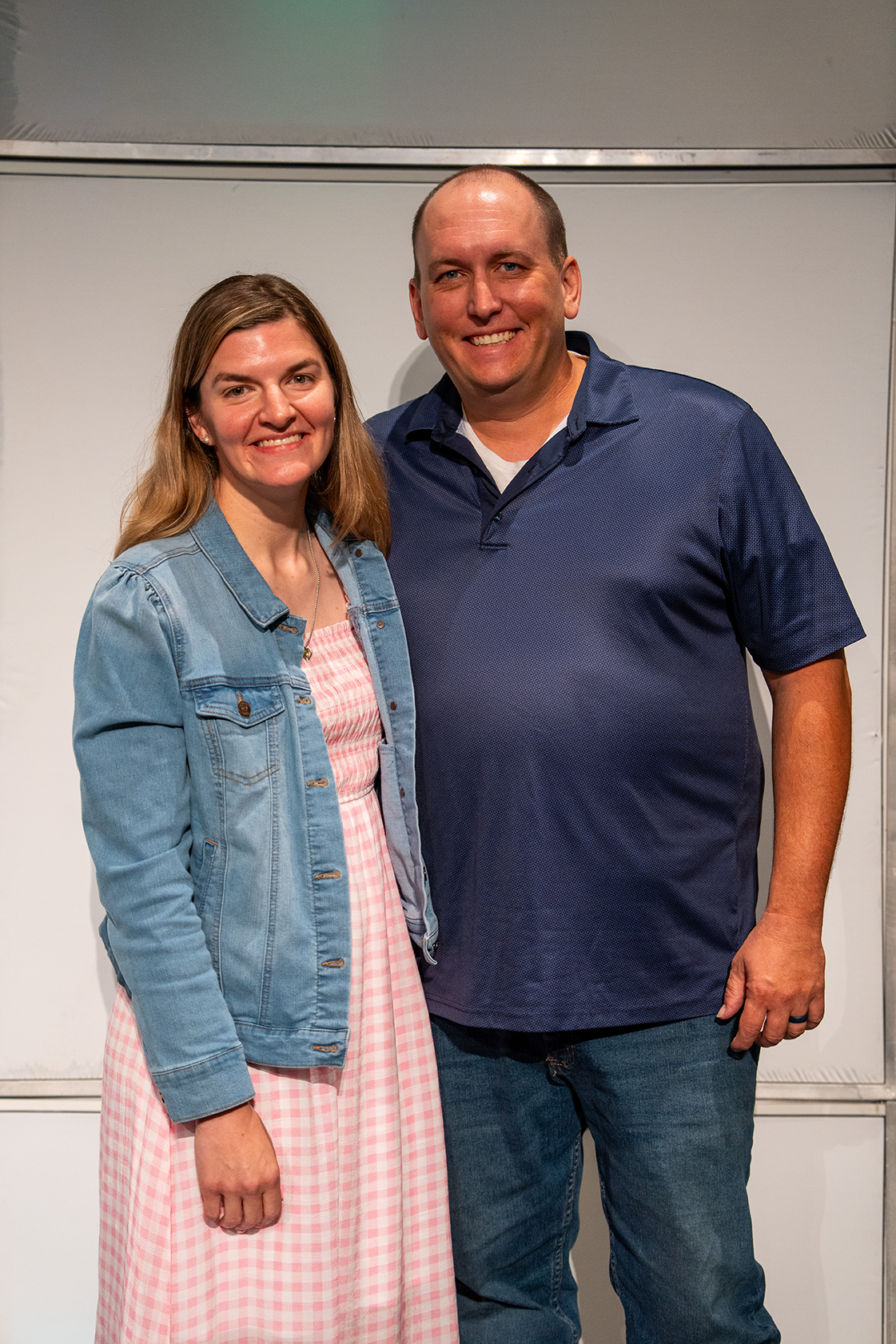 A woman in a pink dress and denim jacket poses next to a man in a navy polo shirt and jeans, both smiling.