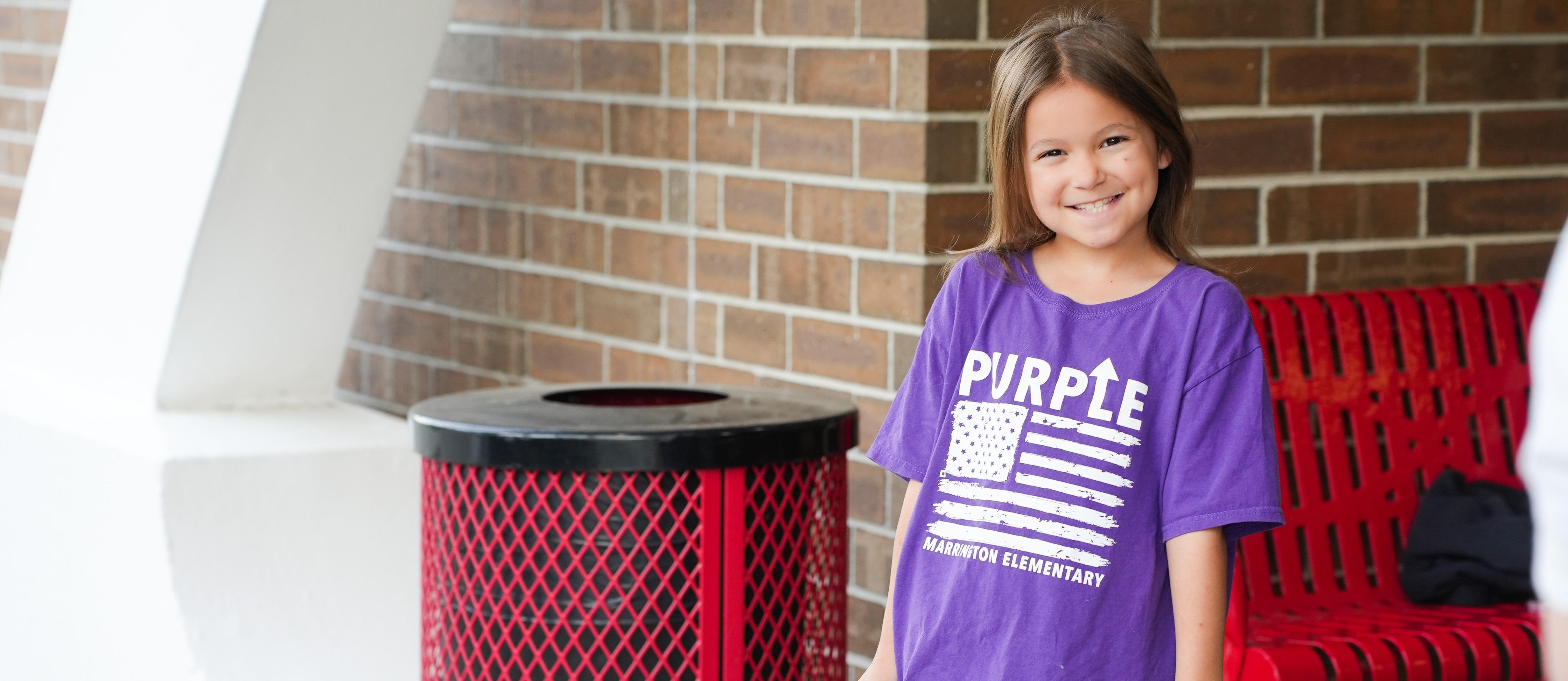A girl in a purple shirt stands near a red bench and trash bin, holding a purple suitcase.
