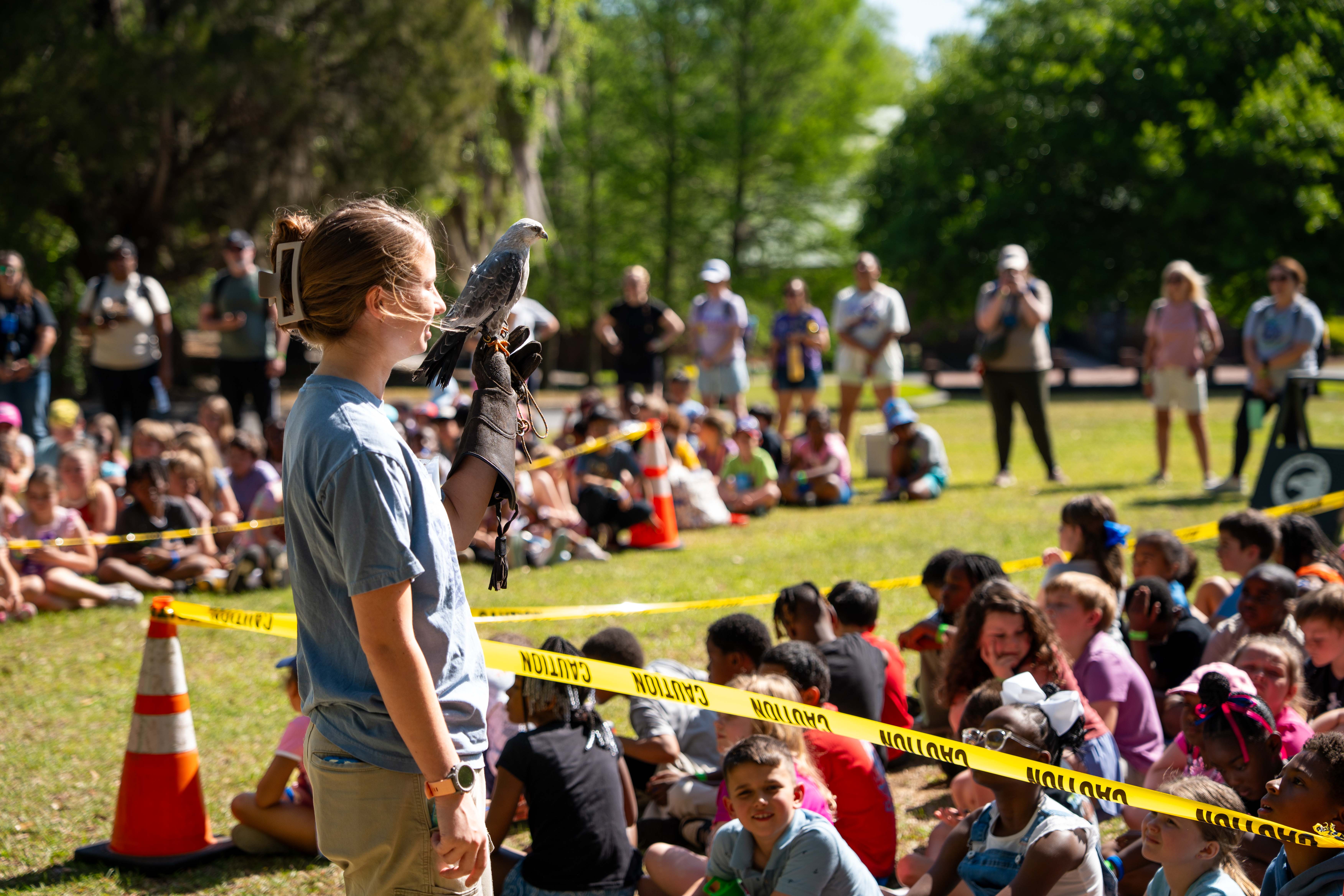 A woman stands with a bird on her arm in a park. Many people sit or stand behind yellow caution tape.