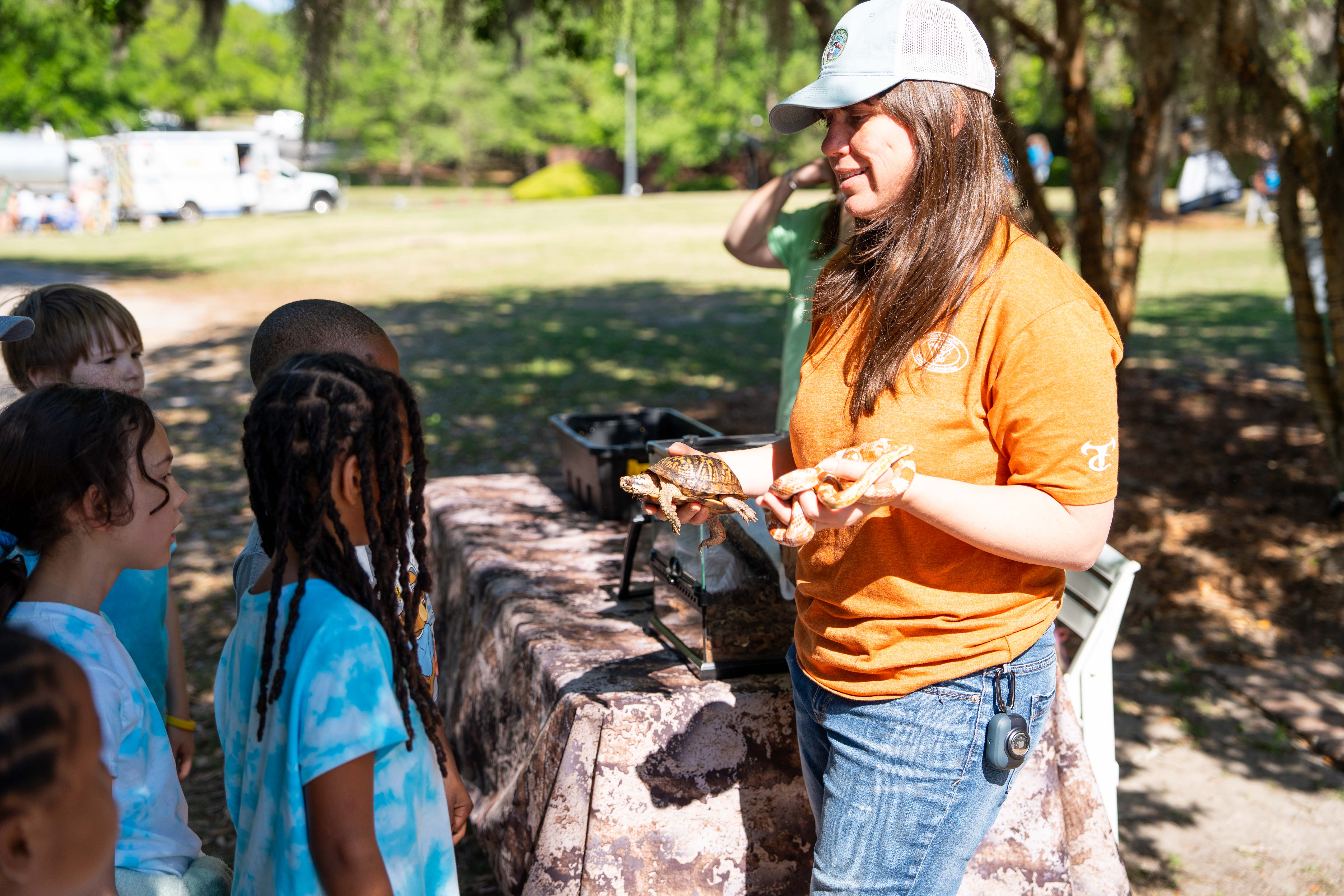 A woman wearing a cap and orange shirt holds a turtle in her hands. Children stand around her.