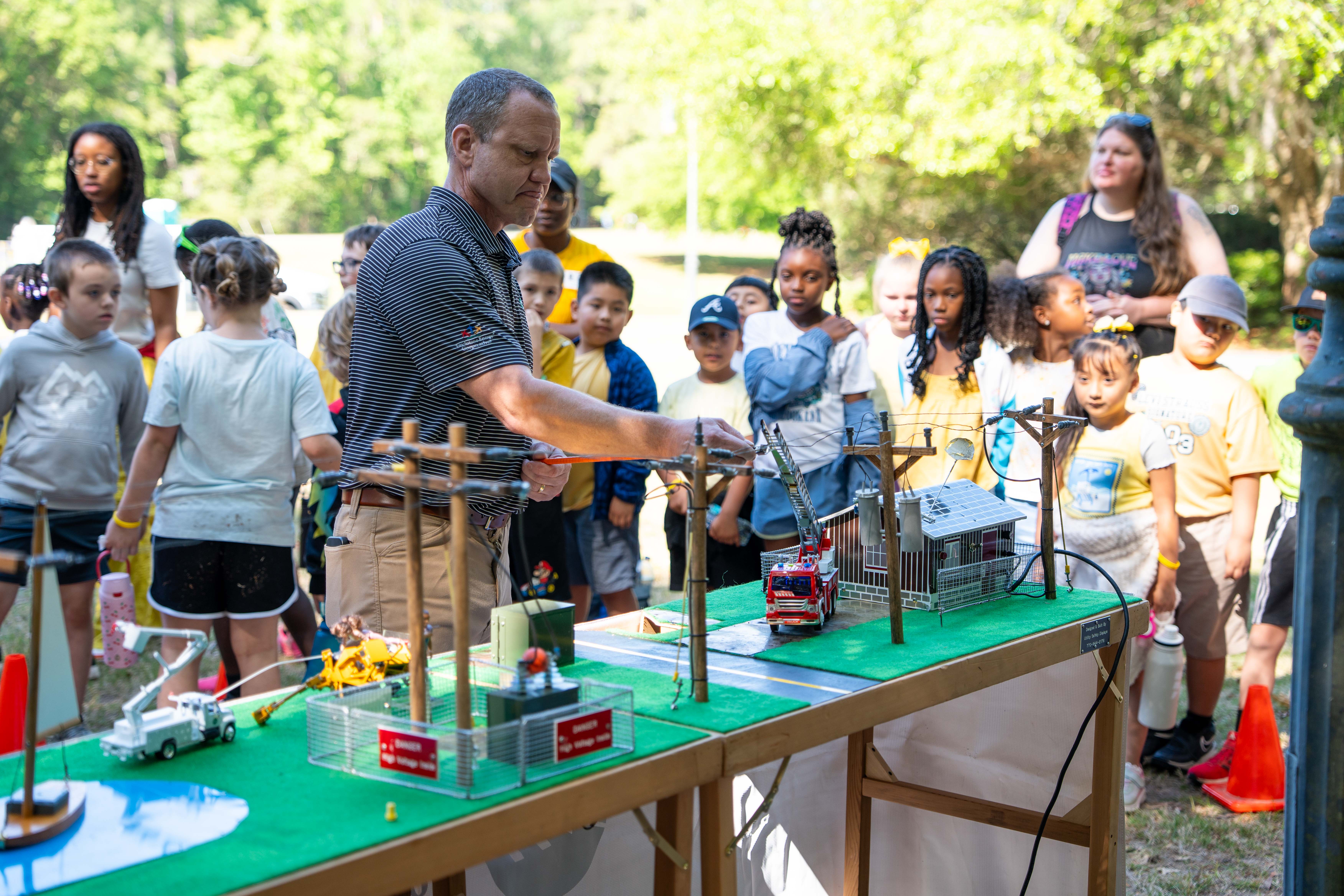 An adult shows a group of children a miniature electrical model on a table in a park.