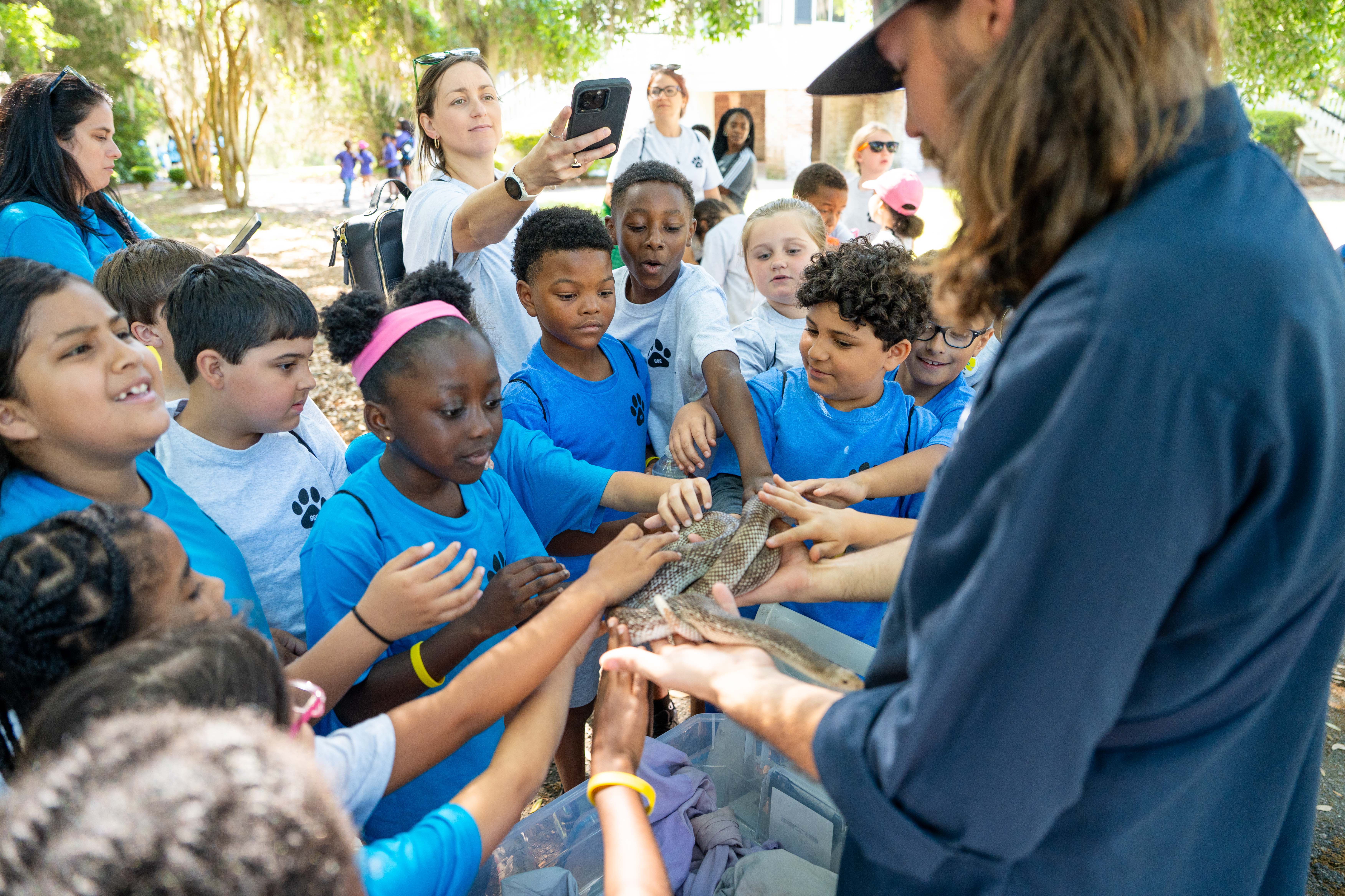 A man in blue holds a reptile for a group of children in blue shirts.