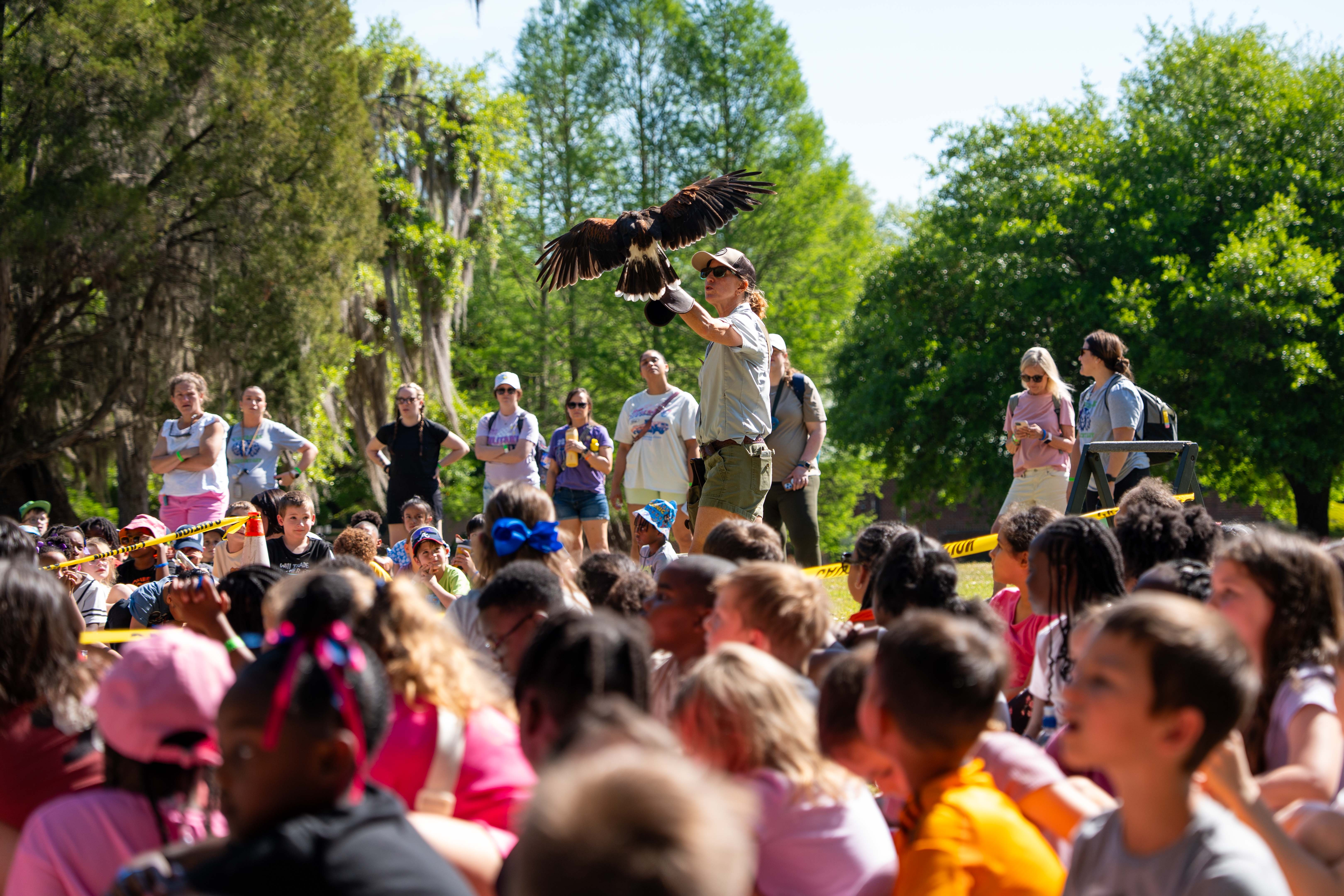 A woman holds a bird over a crowd in a park, with trees and greenery in the background.