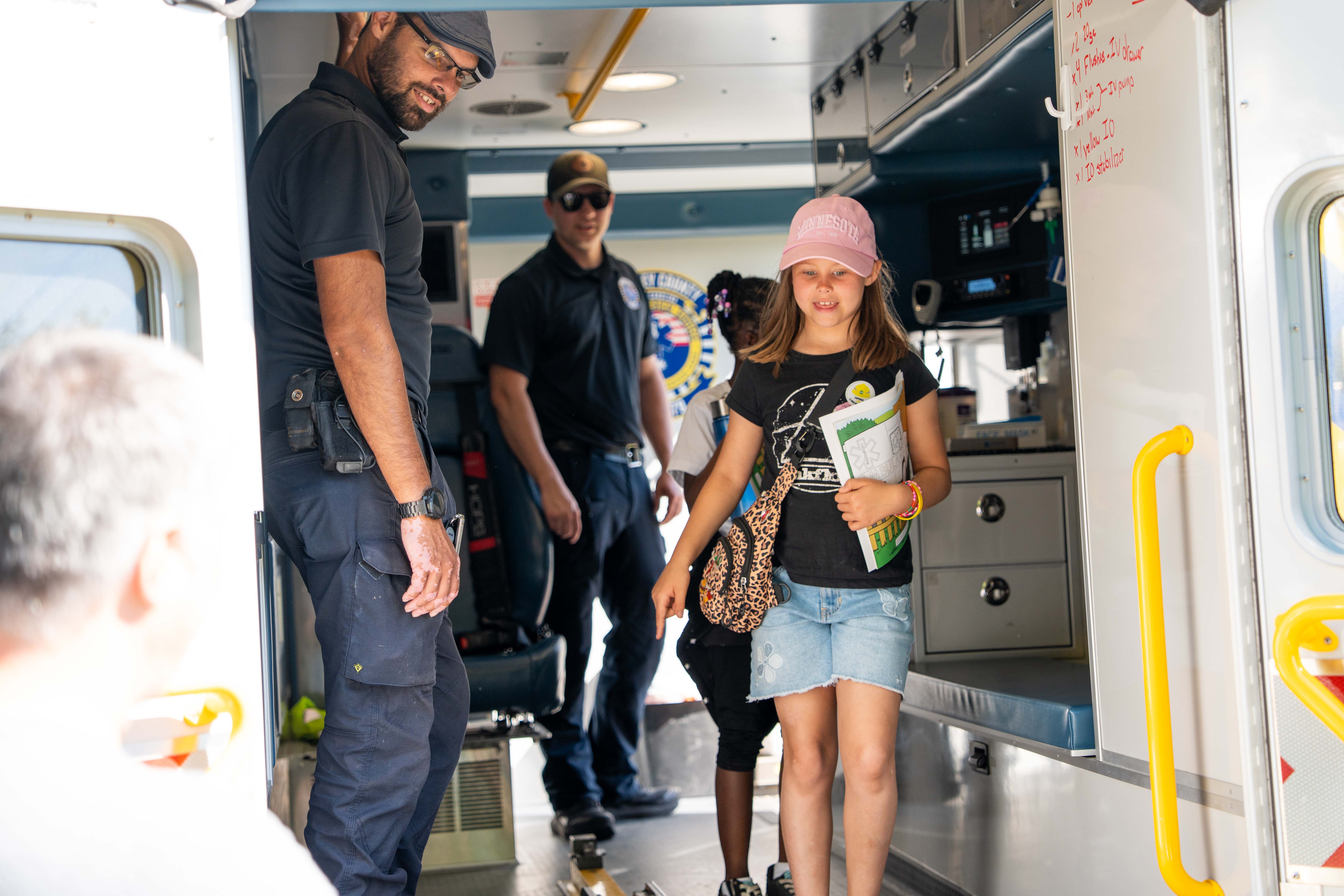 Interior of an ambulance with a young girl in a pink hat and a man in uniform conversing.