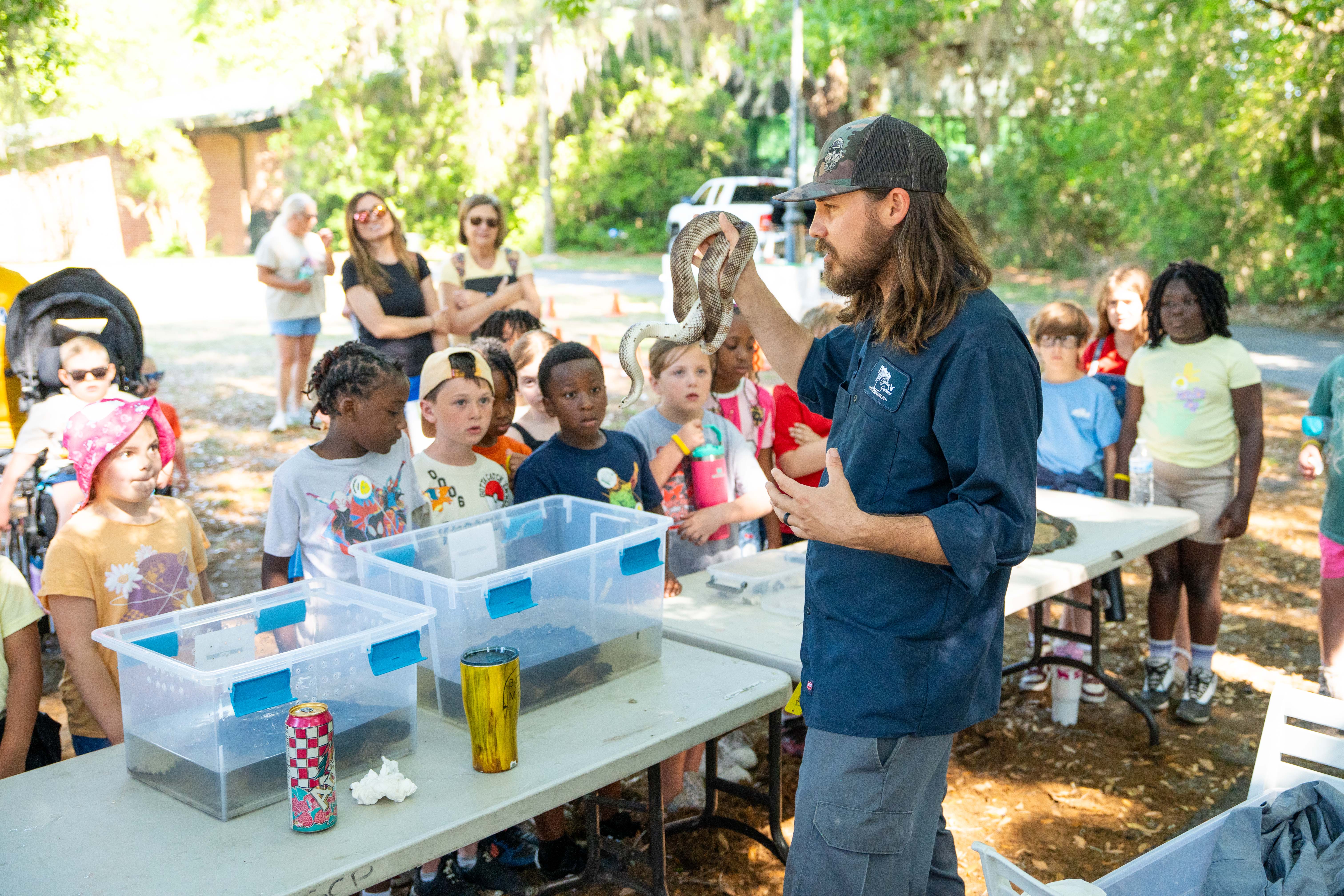 A man holds a snake in a forest area, with children and adults standing behind tables and chairs.