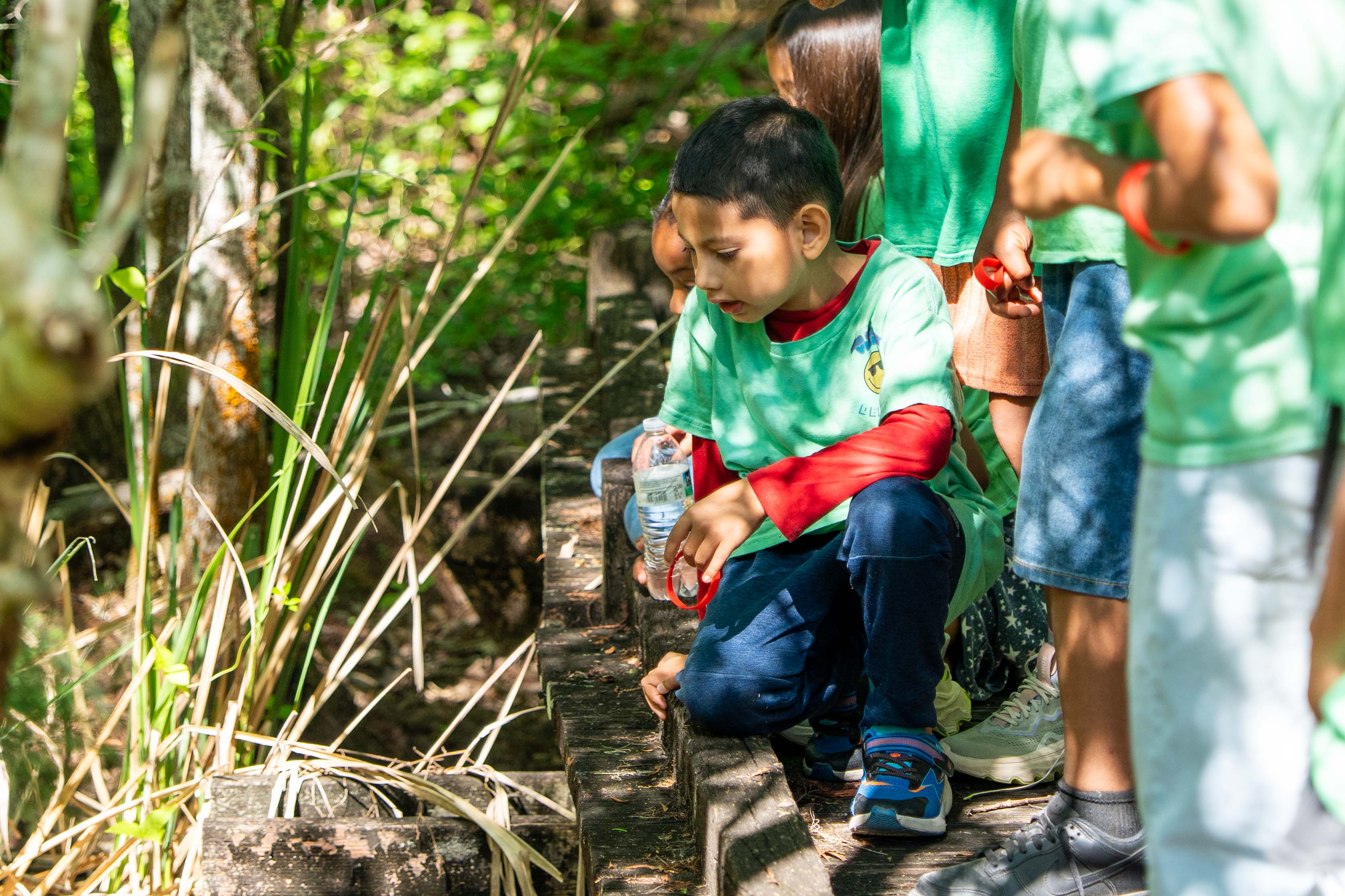 Children in green shirts, one kneeling with a bottle, another standing on a forest path.