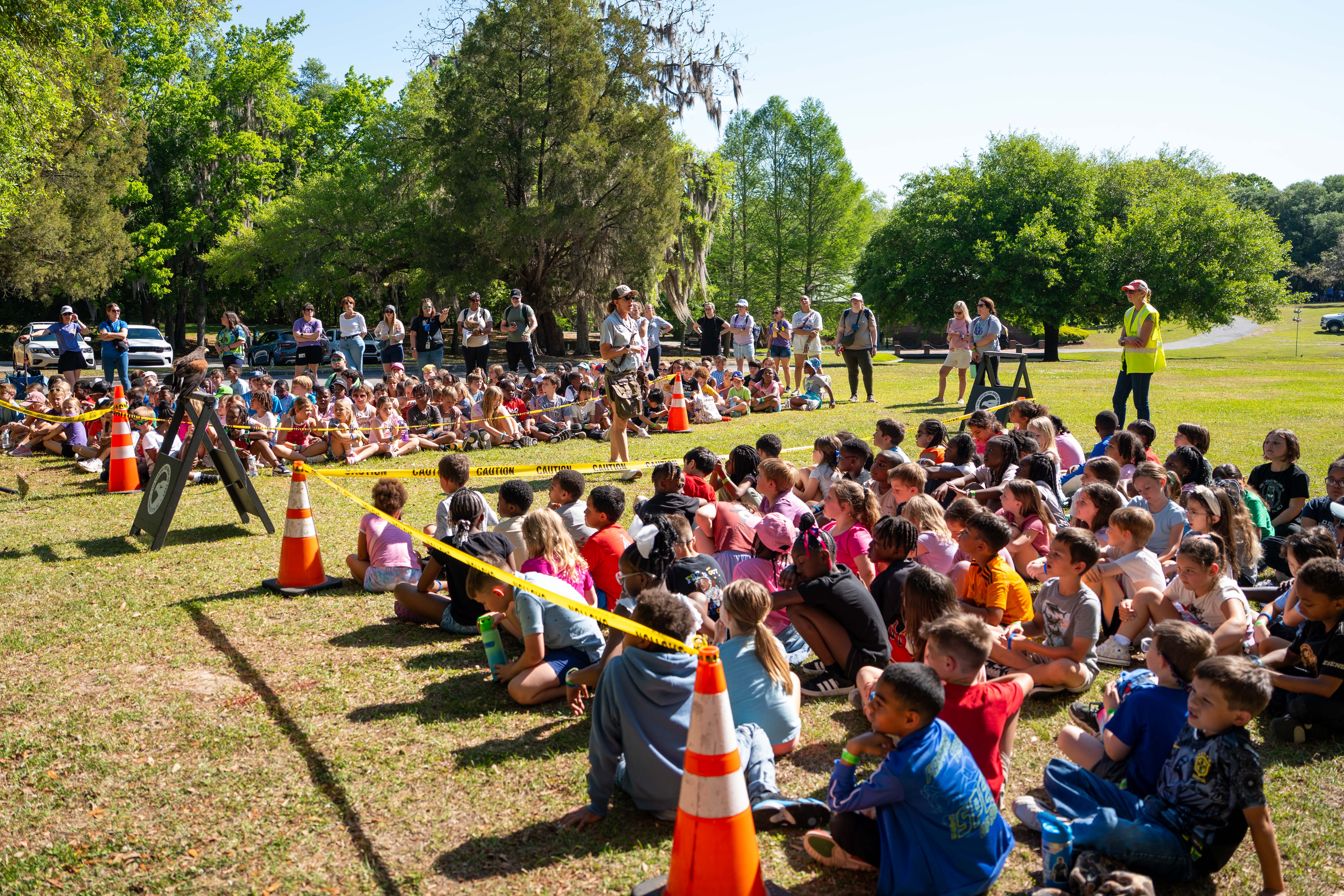 Many children sit on grass in a park. A yellow line blocks part of the area. People stand and sit at the back.