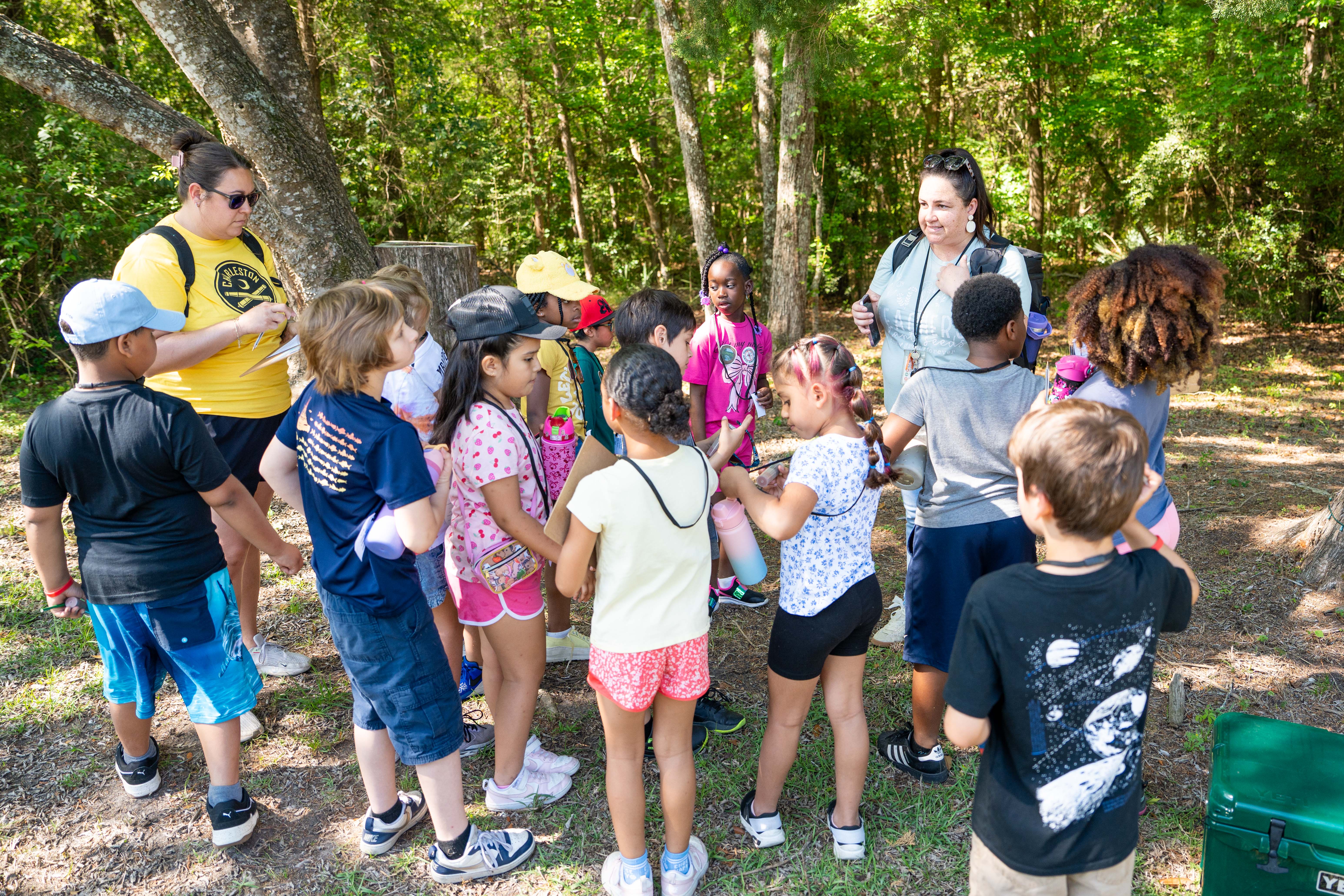 A group of children, some wearing hats, sneakers, and sunglasses, stands in a circle outdoors with trees in the background.