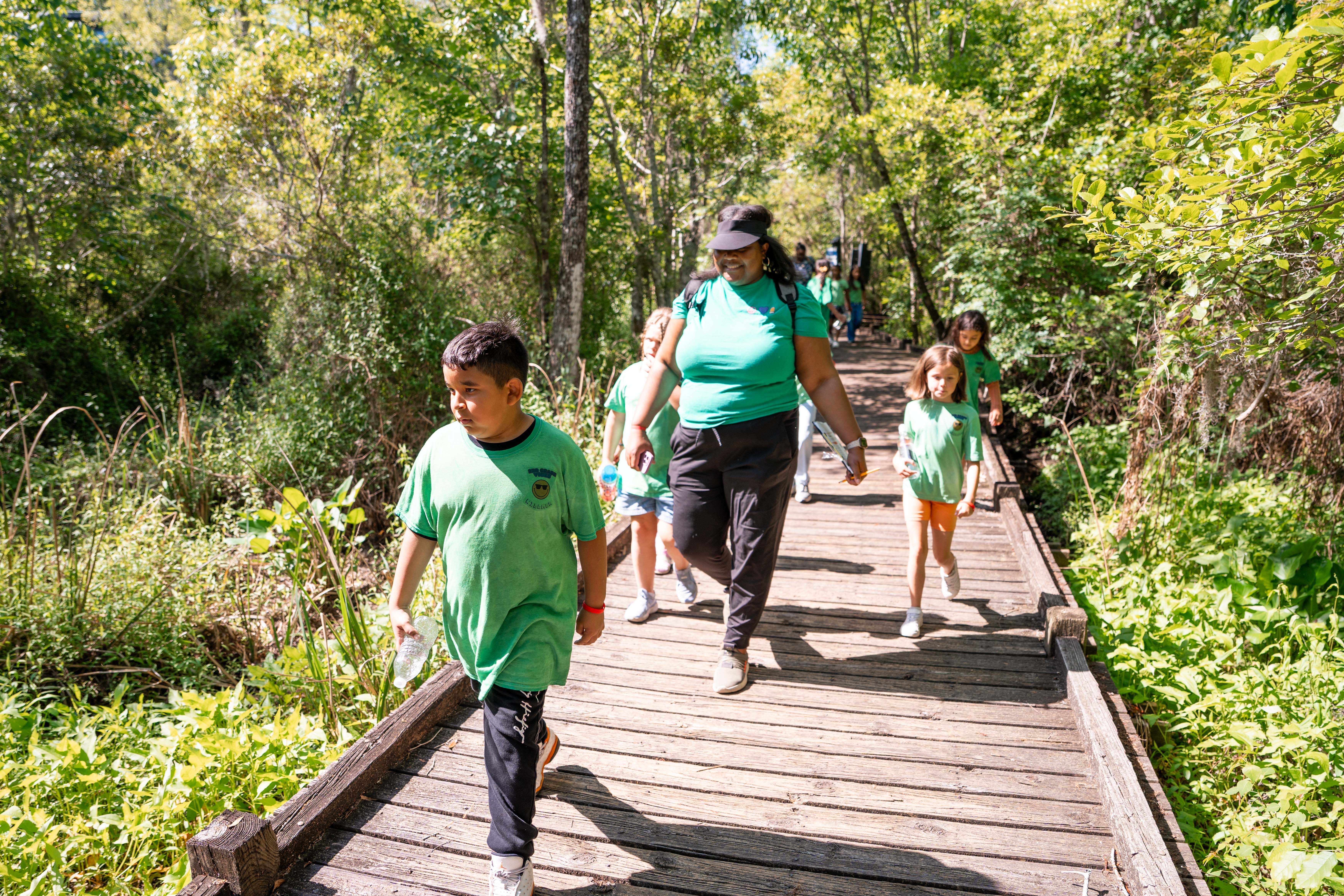 A group of children and adults wearing green shirts walks on a wooden path through a forest.