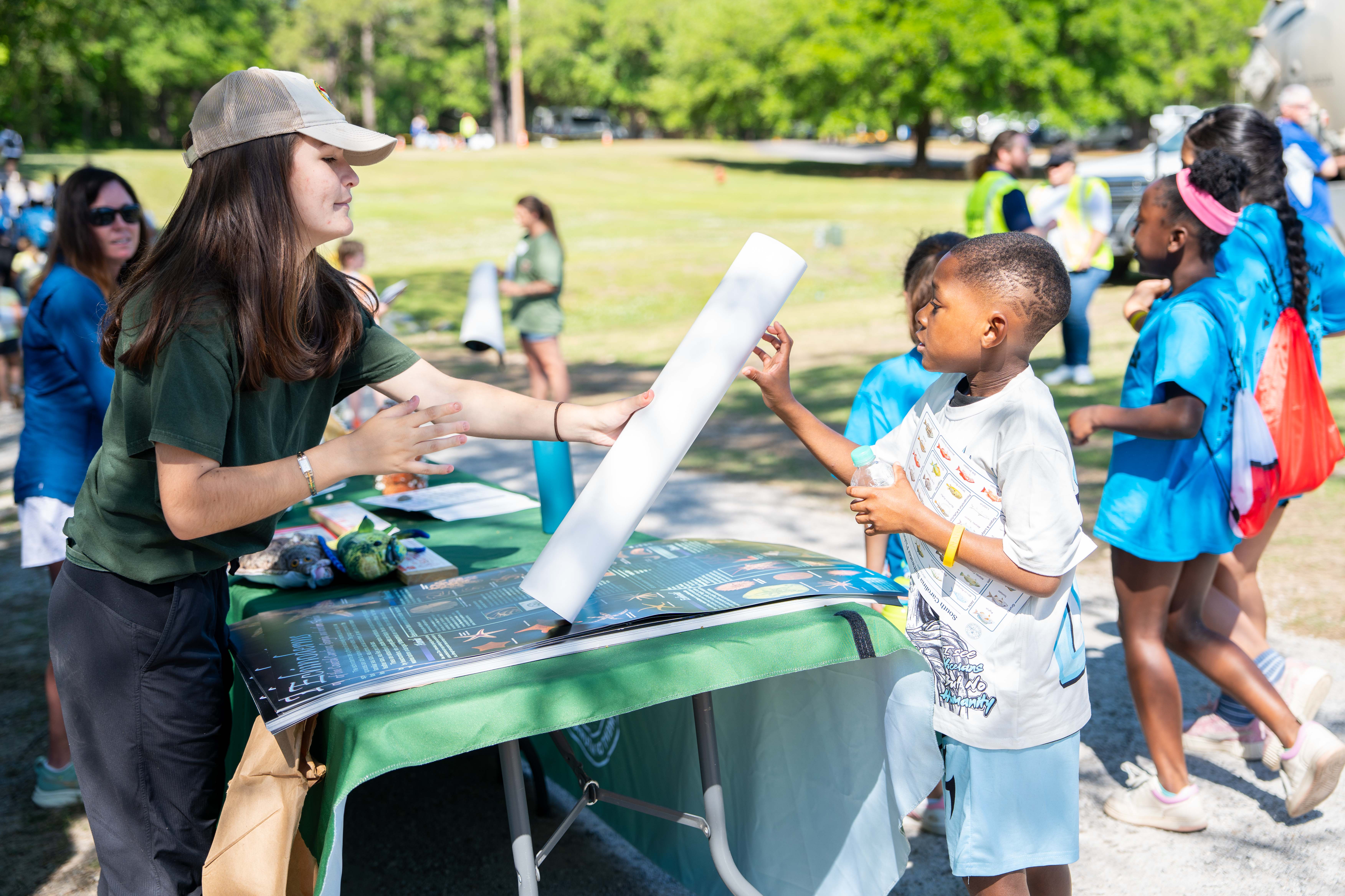 Woman in green shirt holds a rolled paper in front of a boy at an outdoor event.