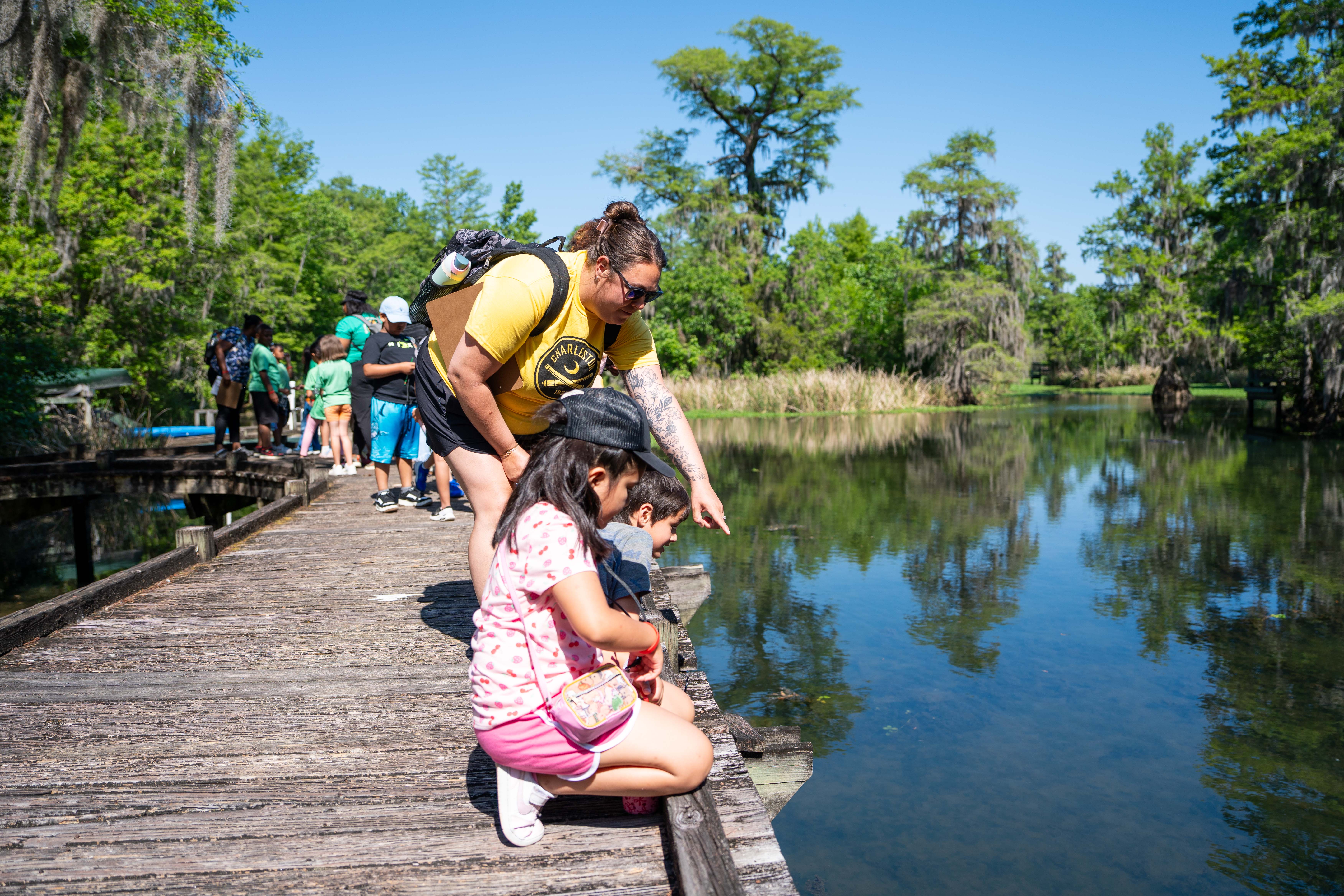 A woman and a child crouch by a pond on a wooden dock with trees in the background.