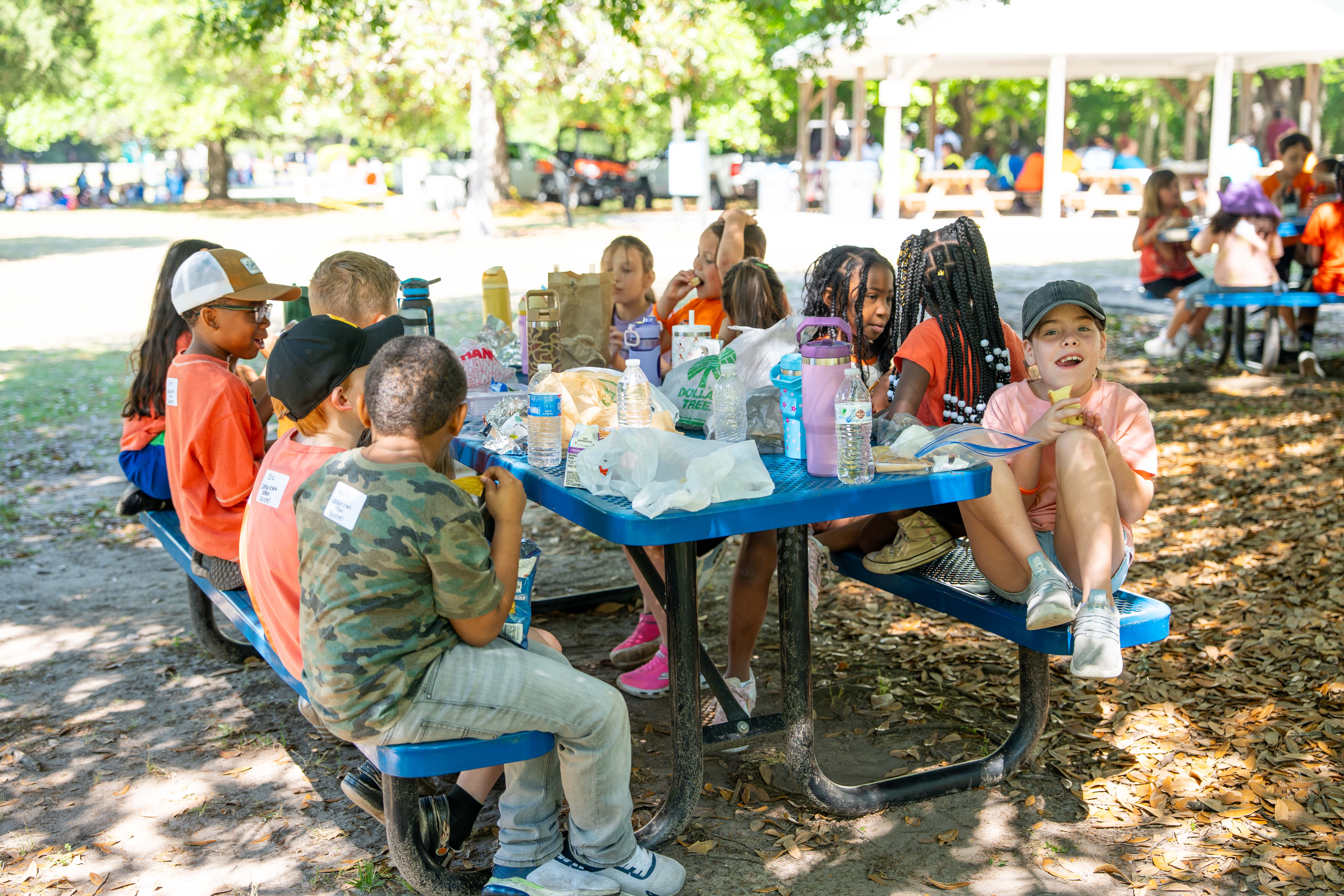 Children seated at a picnic table in a park, with others sitting at tables and a shelter in the background.