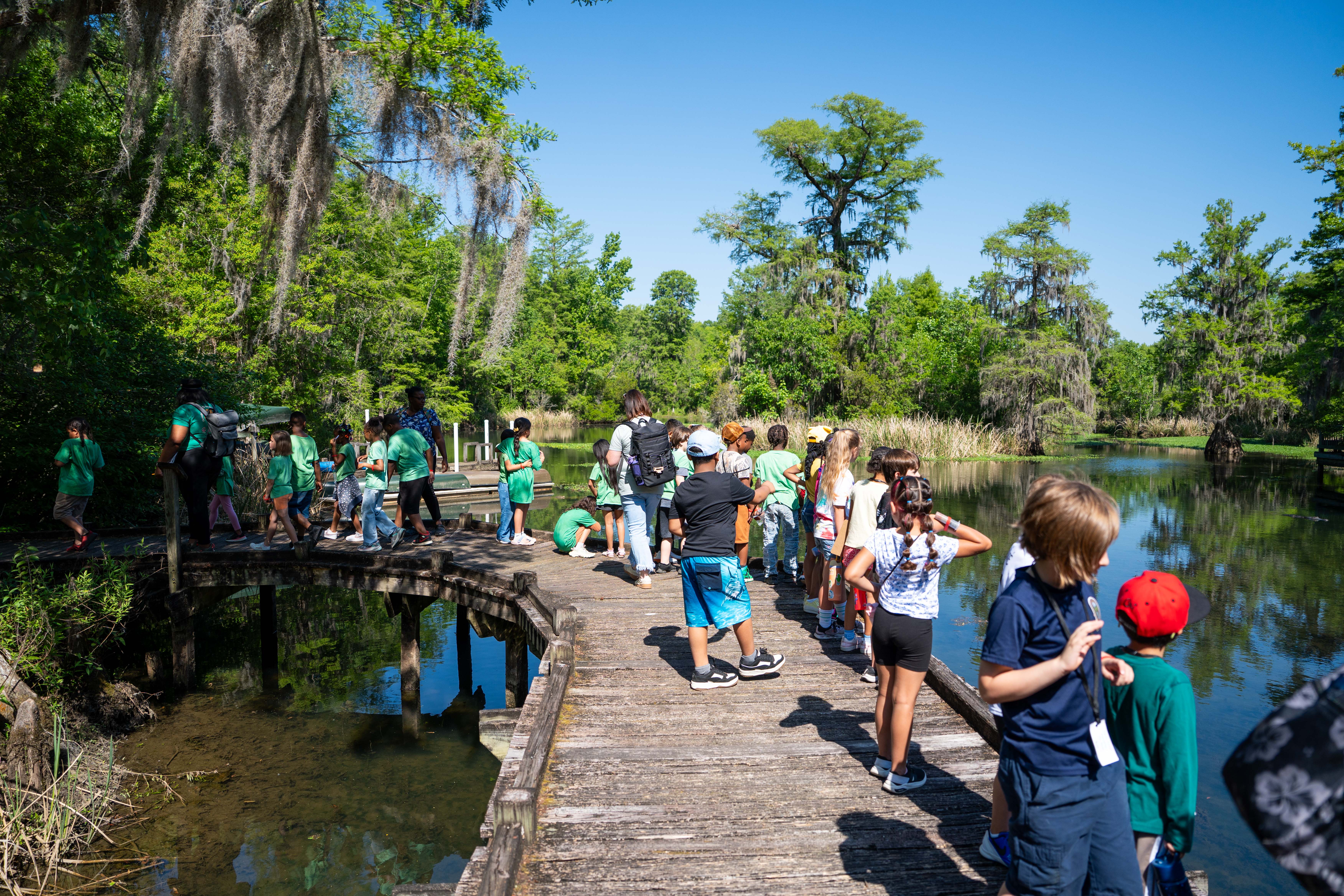 A group of people on a wooden dock by a pond, wearing green shirts, hats, and sneakers.