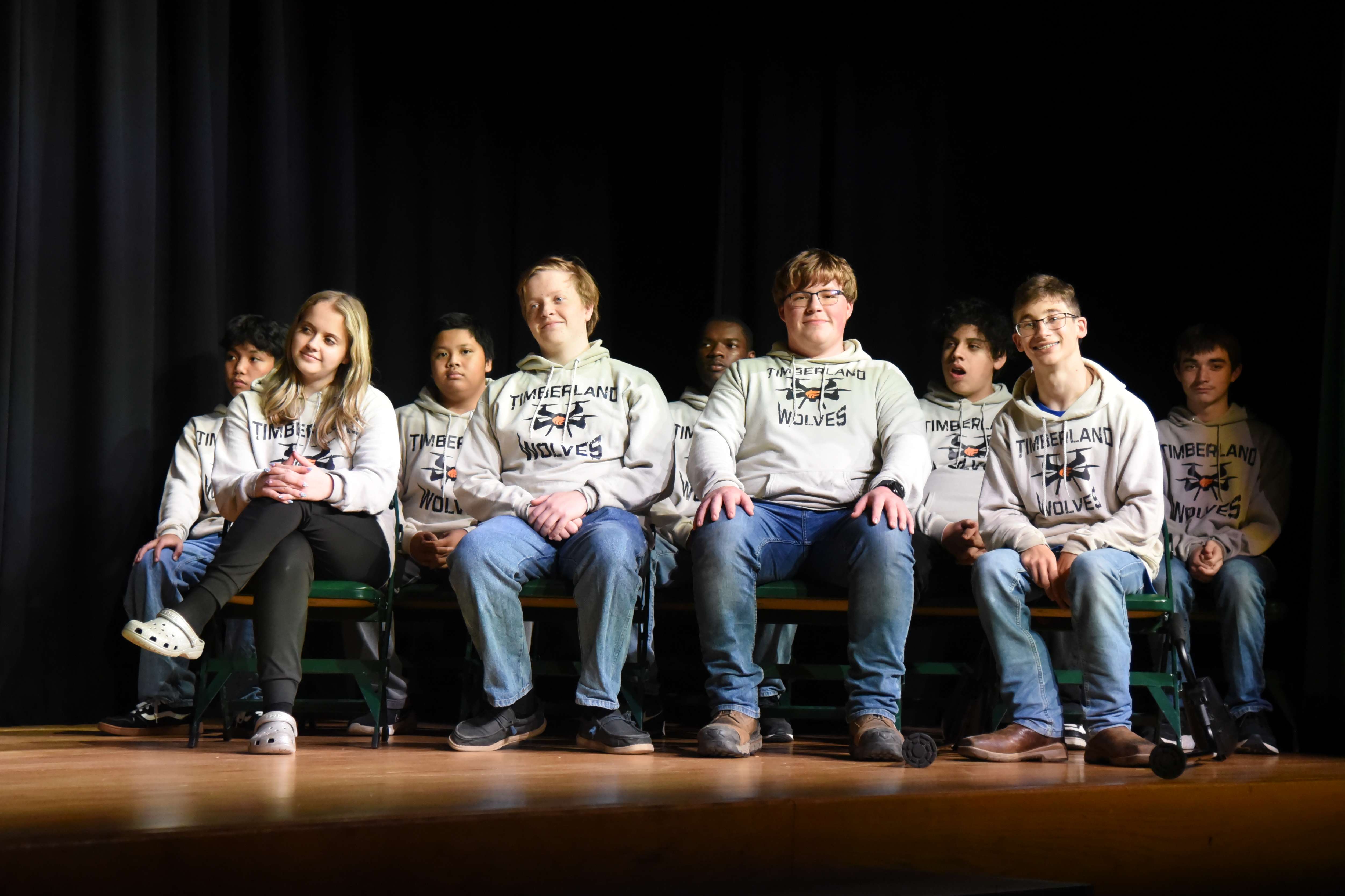 A group of people in matching white hoodies and jeans seated on a bench on a stage.