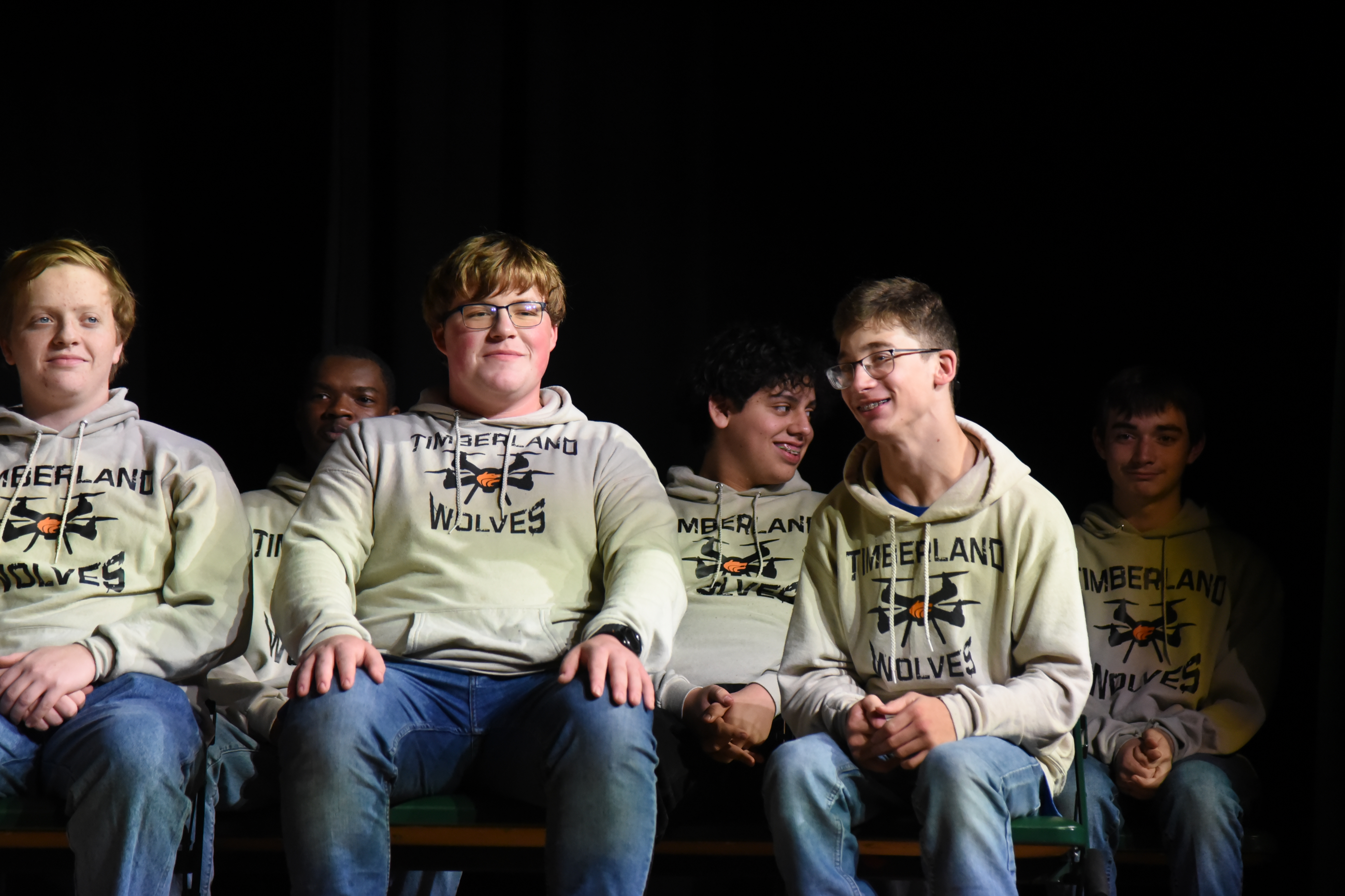 Five boys in sweatshirts sit together on a bench, facing the camera. Three of them wear glasses.
