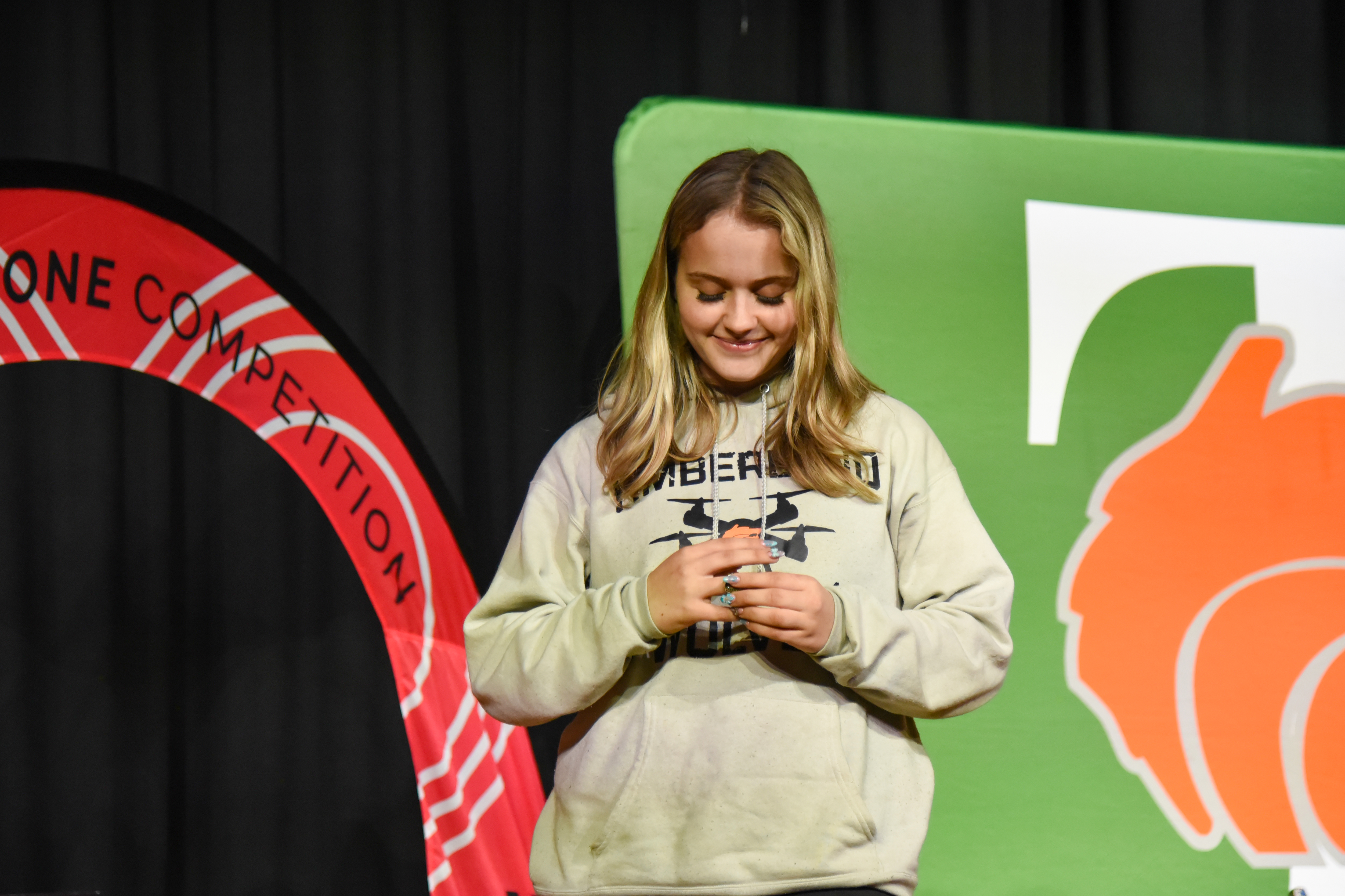 A woman on stage wearing a white sweatshirt, smiling, and holding something in her hands. Behind her are banners.