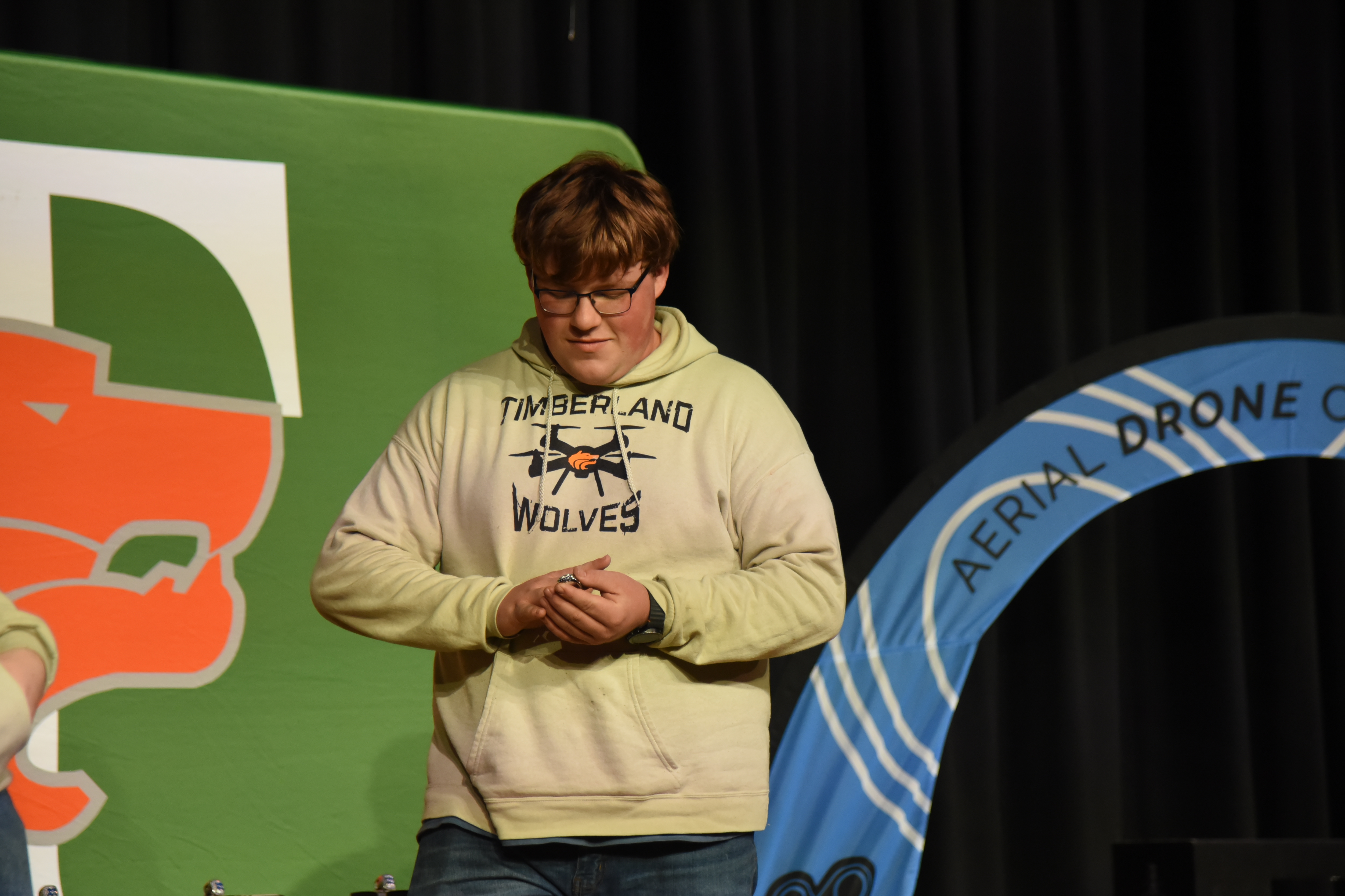 A person in a light yellow hoodie with a black print stands before a stage backdrop with a blue banner.