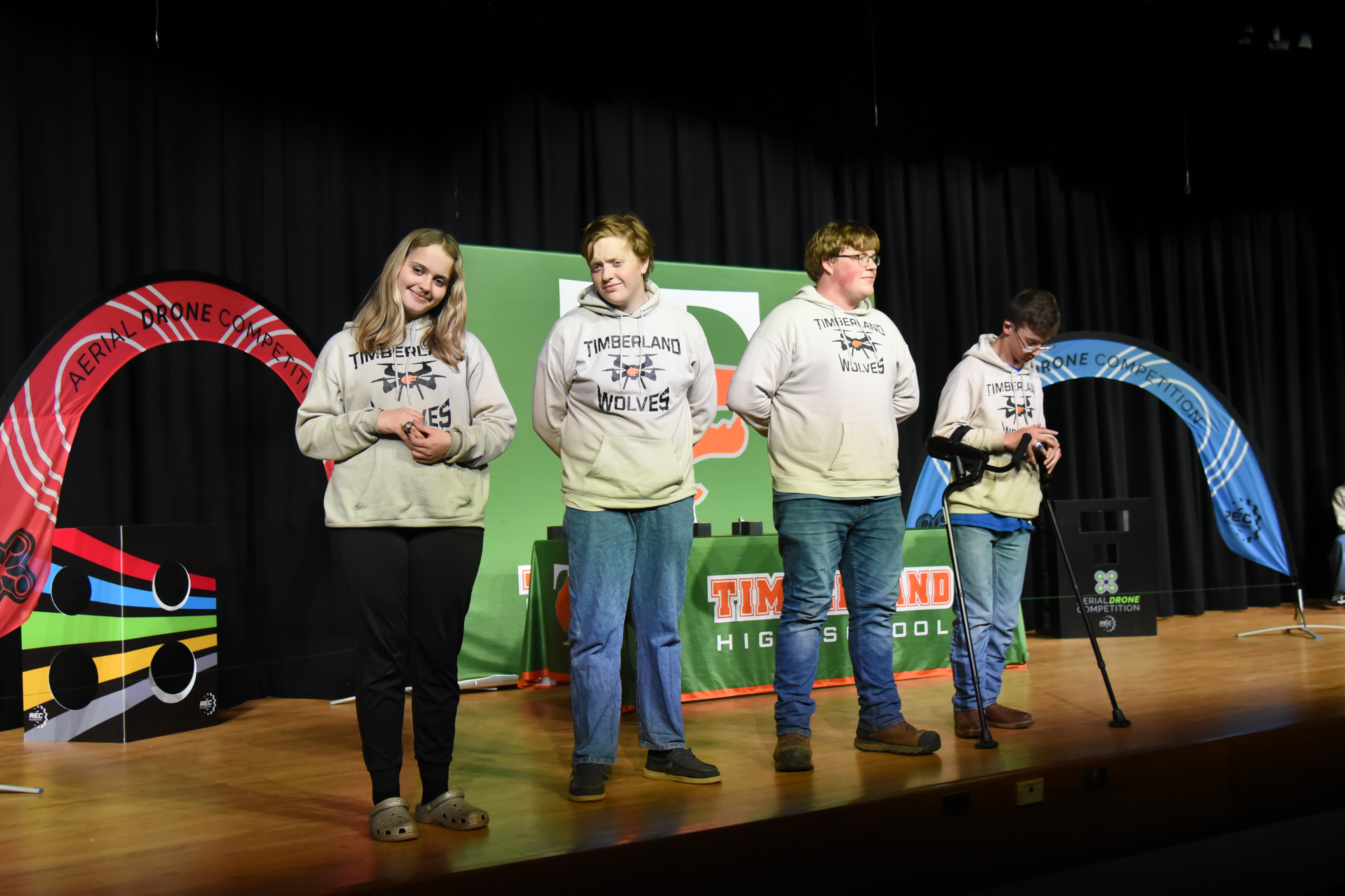 Four people in hoodies and pants stand on a stage, next to a colorful arch. One person holds a microphone.