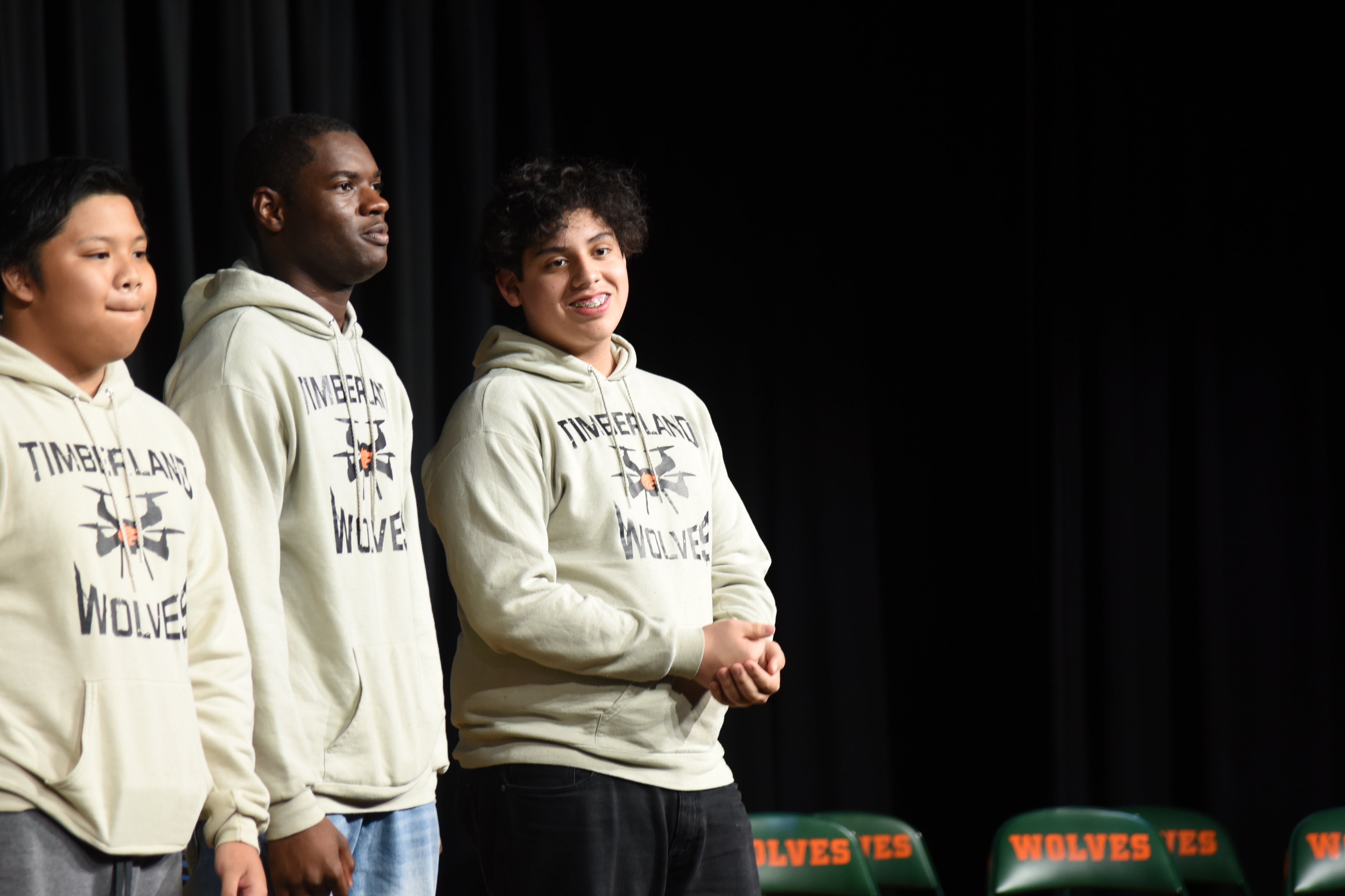Three people in white hoodies with a black background and green chairs. One person is wearing a white sweatshirt.