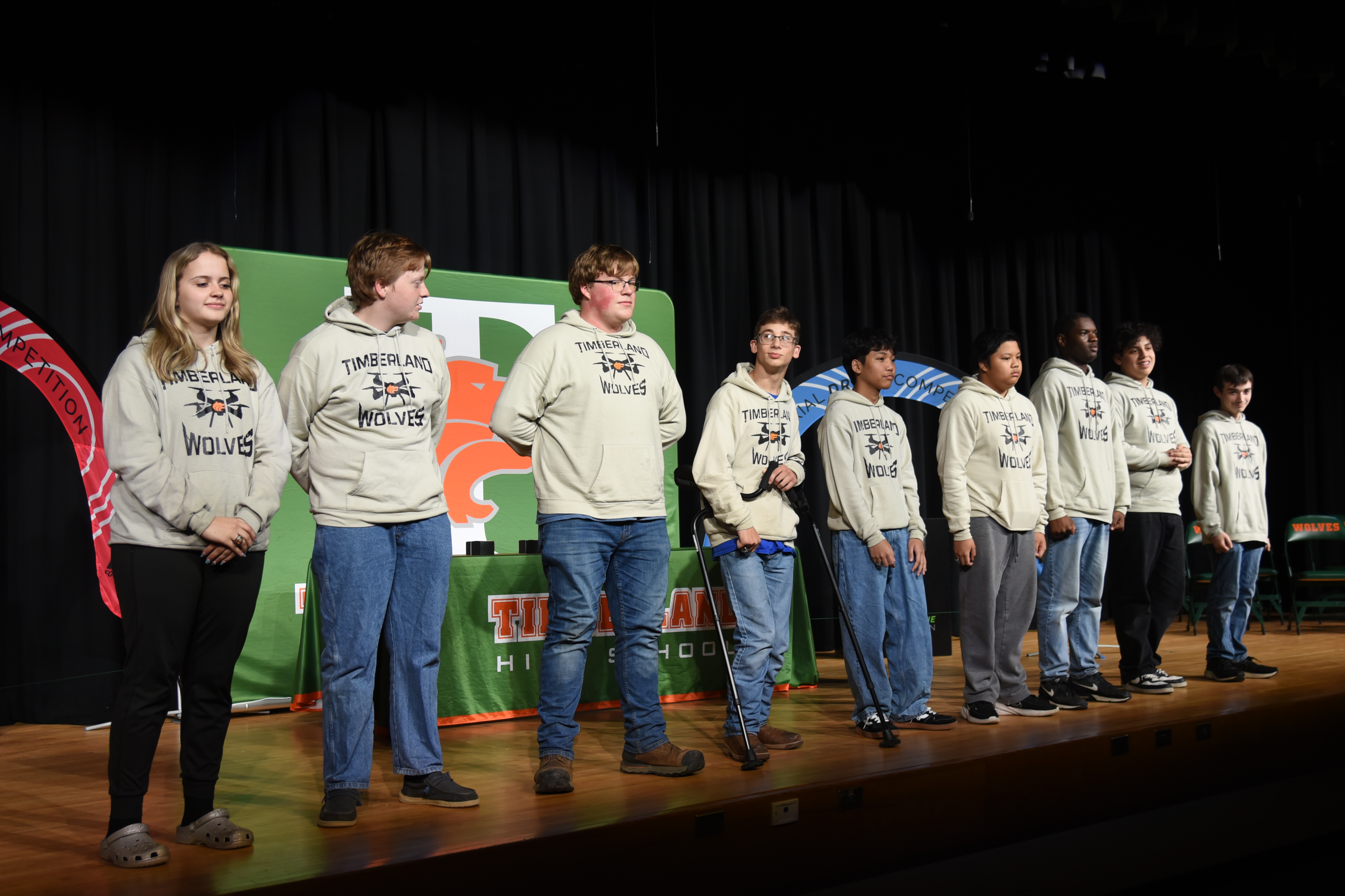 Seven students stand on a stage, wearing matching sweatshirts. A banner behind them displays an image.