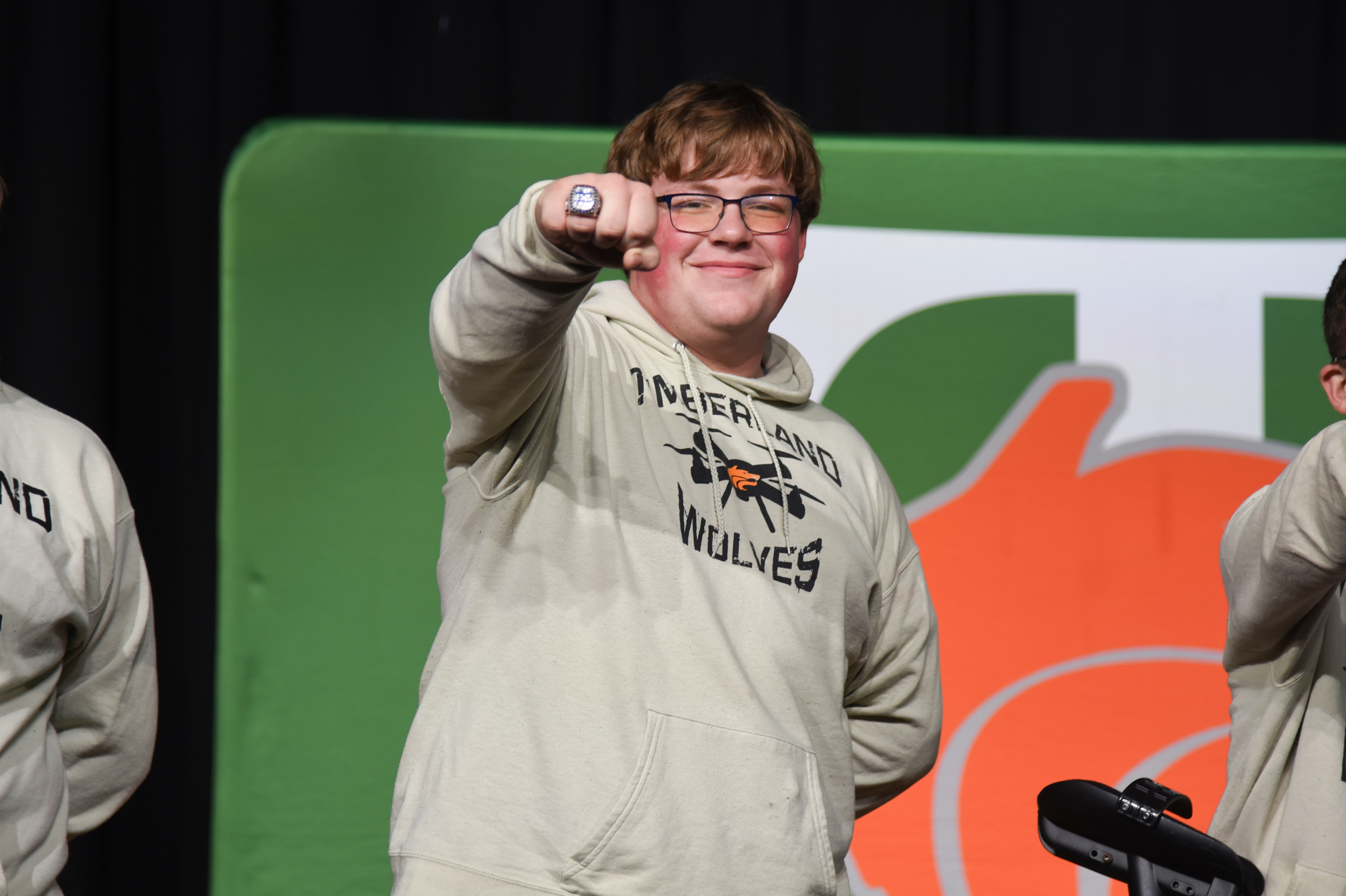 Person in gray hoodie smiles and gestures with ring. Two others in white hoodies stand nearby. Green and orange backdrop behind.