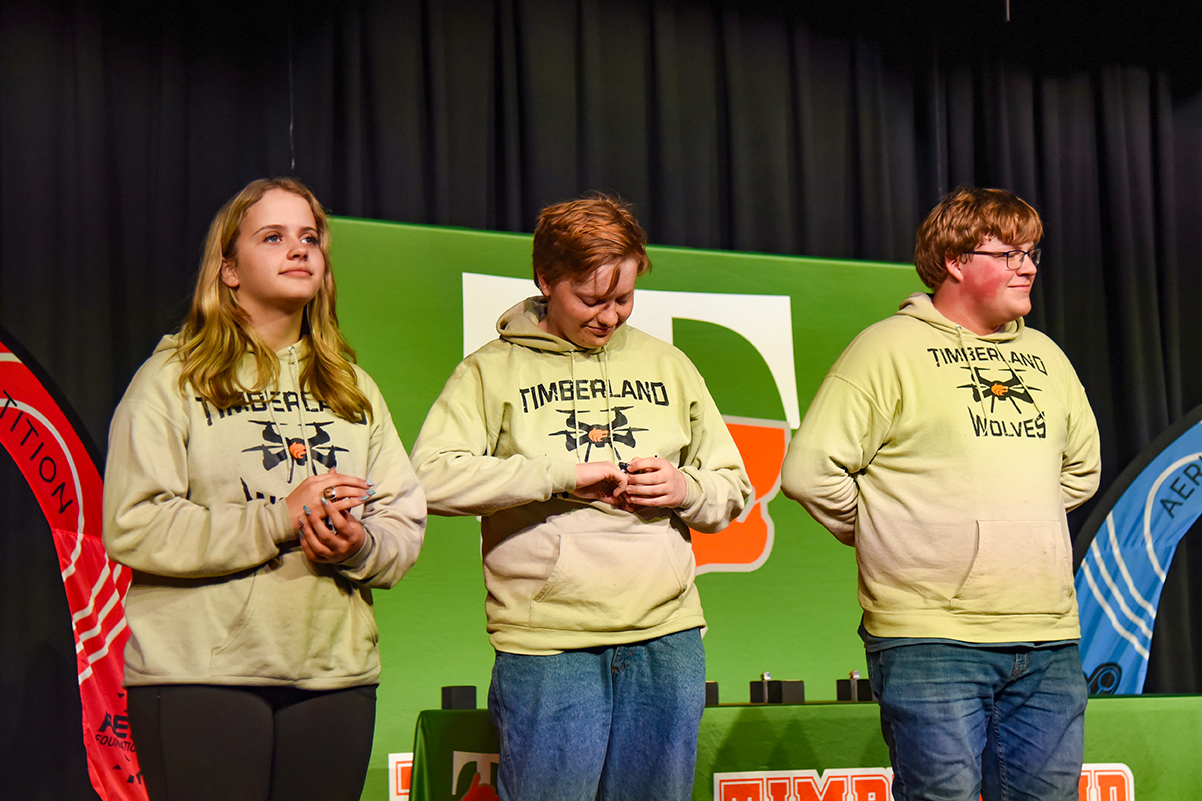 Three young adults, one female and two males, in yellow sweatshirts stand on a stage with a green backdrop.