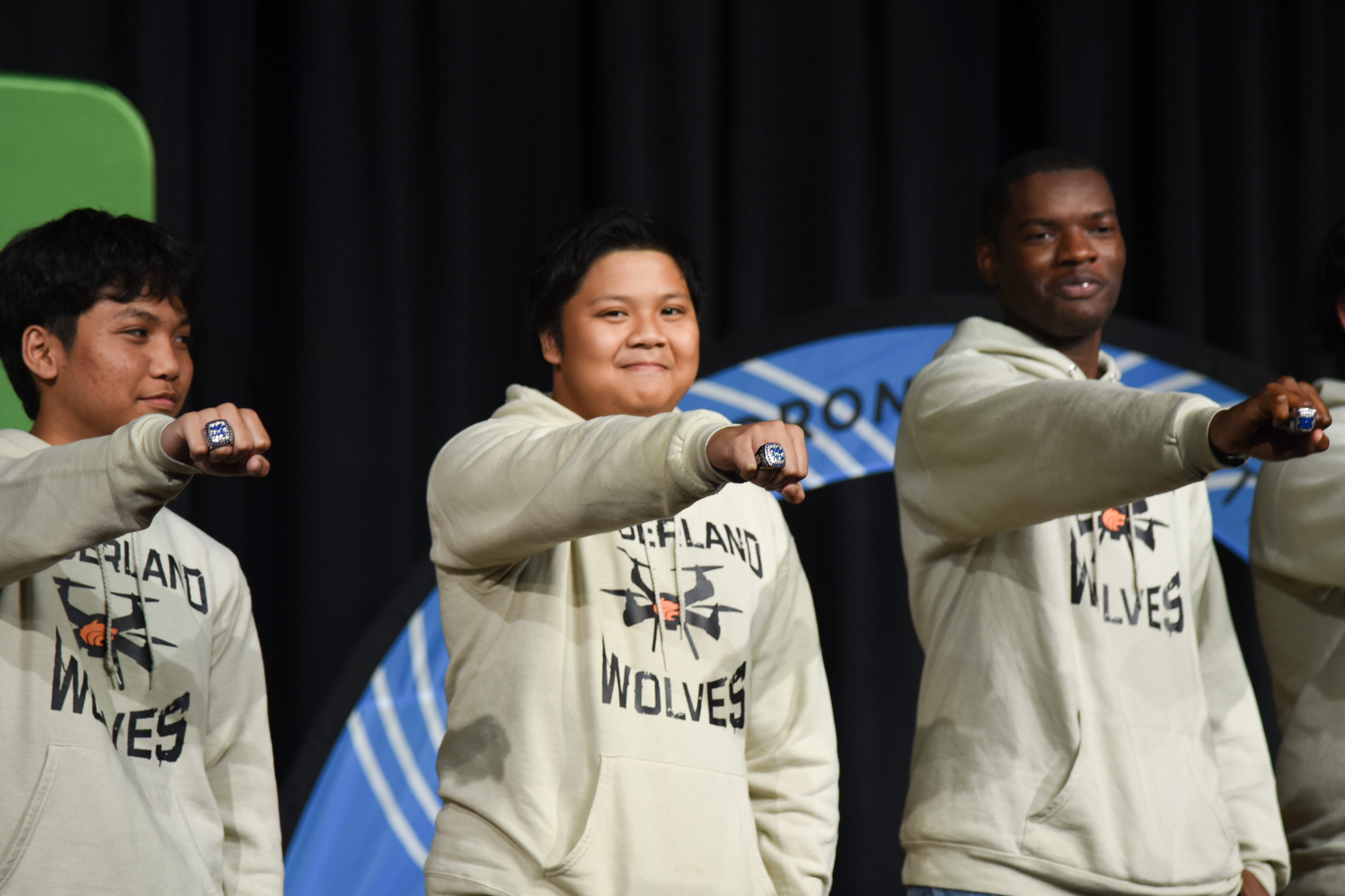Four people in matching sweatshirts smile while performing sign language on a stage. A blue circle with text is behind them.