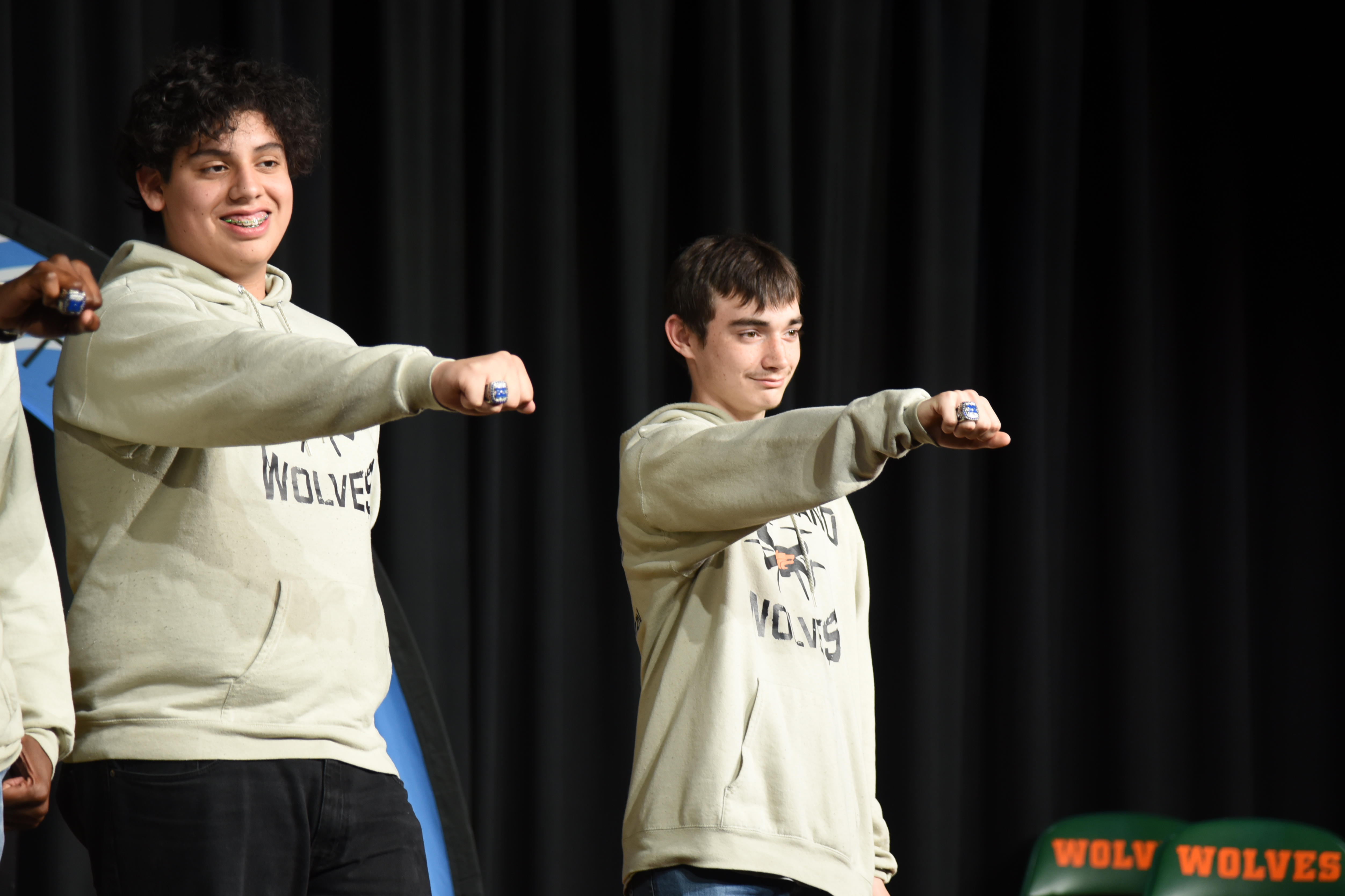 Two young men in light-colored sweatshirts and black pants are performing on stage with hands extended.