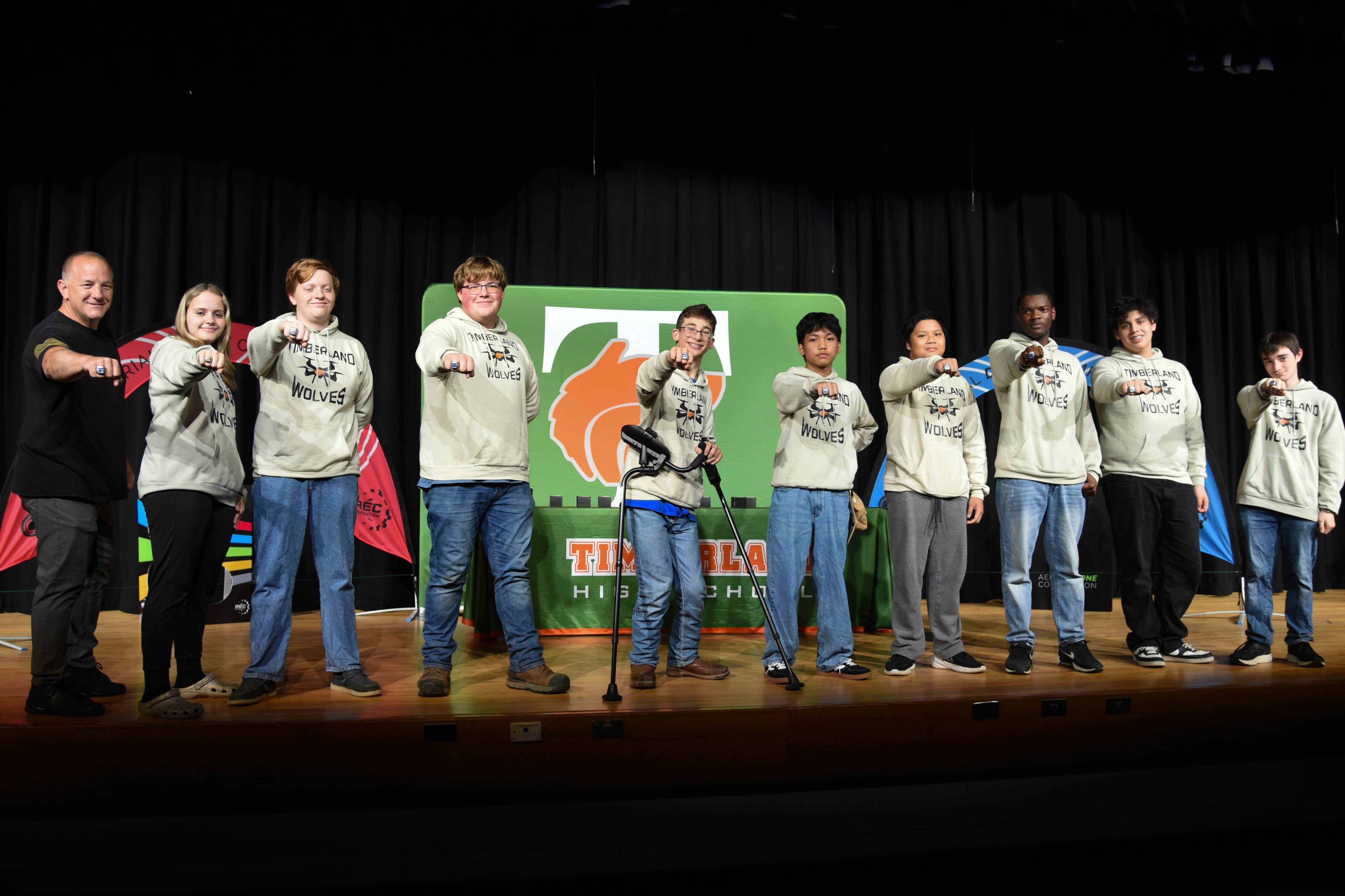 Nine individuals wearing matching light-colored hoodies stand on a stage, some with hands raised, a banner behind them.