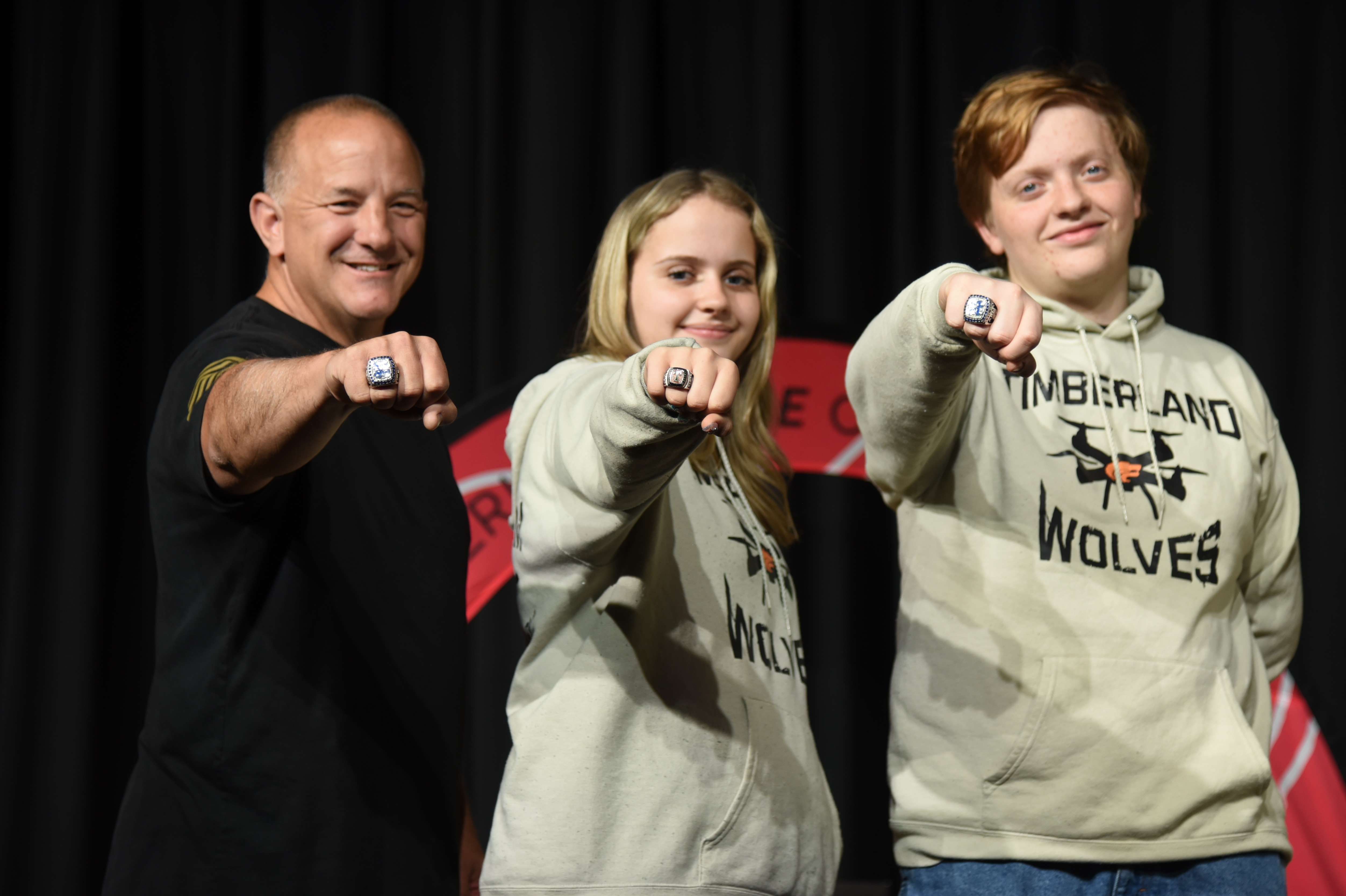 Three individuals pose with arms raised. The man in black, two women in gray hoodies. Background includes a red and black banner.