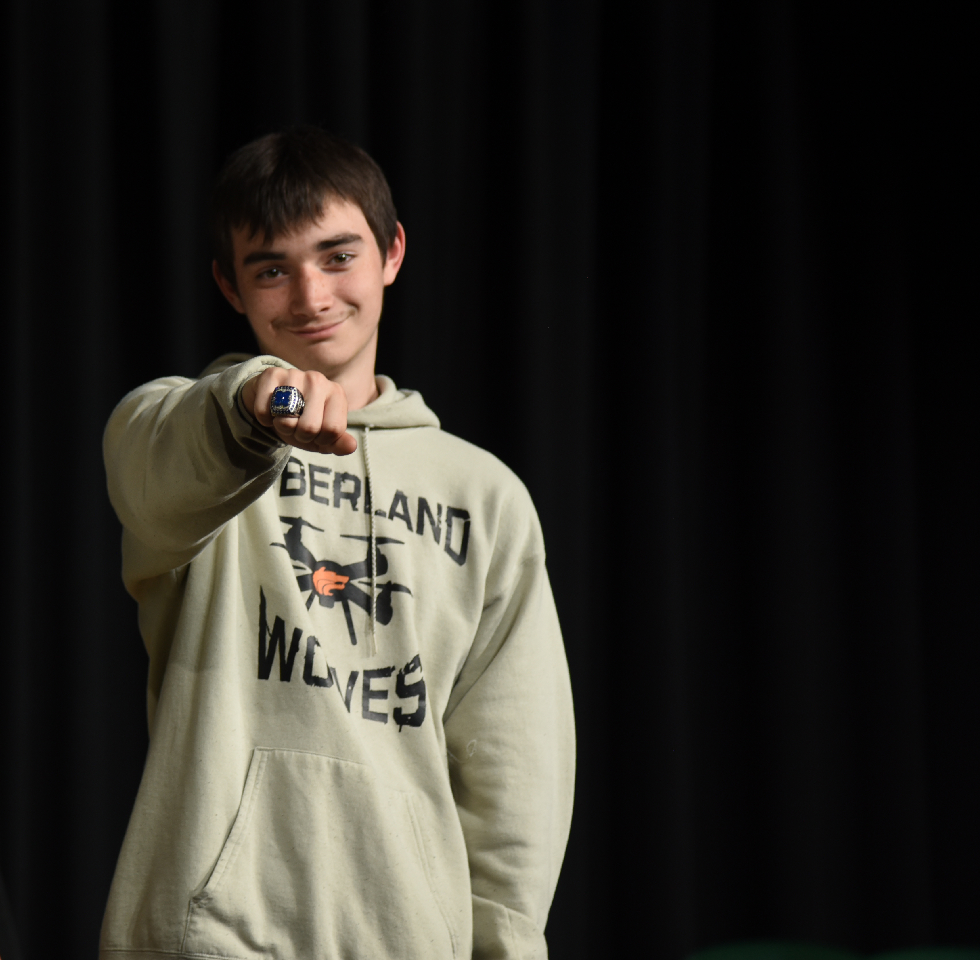 A person wearing a light-colored hoodie with "BERLAND WOLVES" and a drone logo smiles and points.