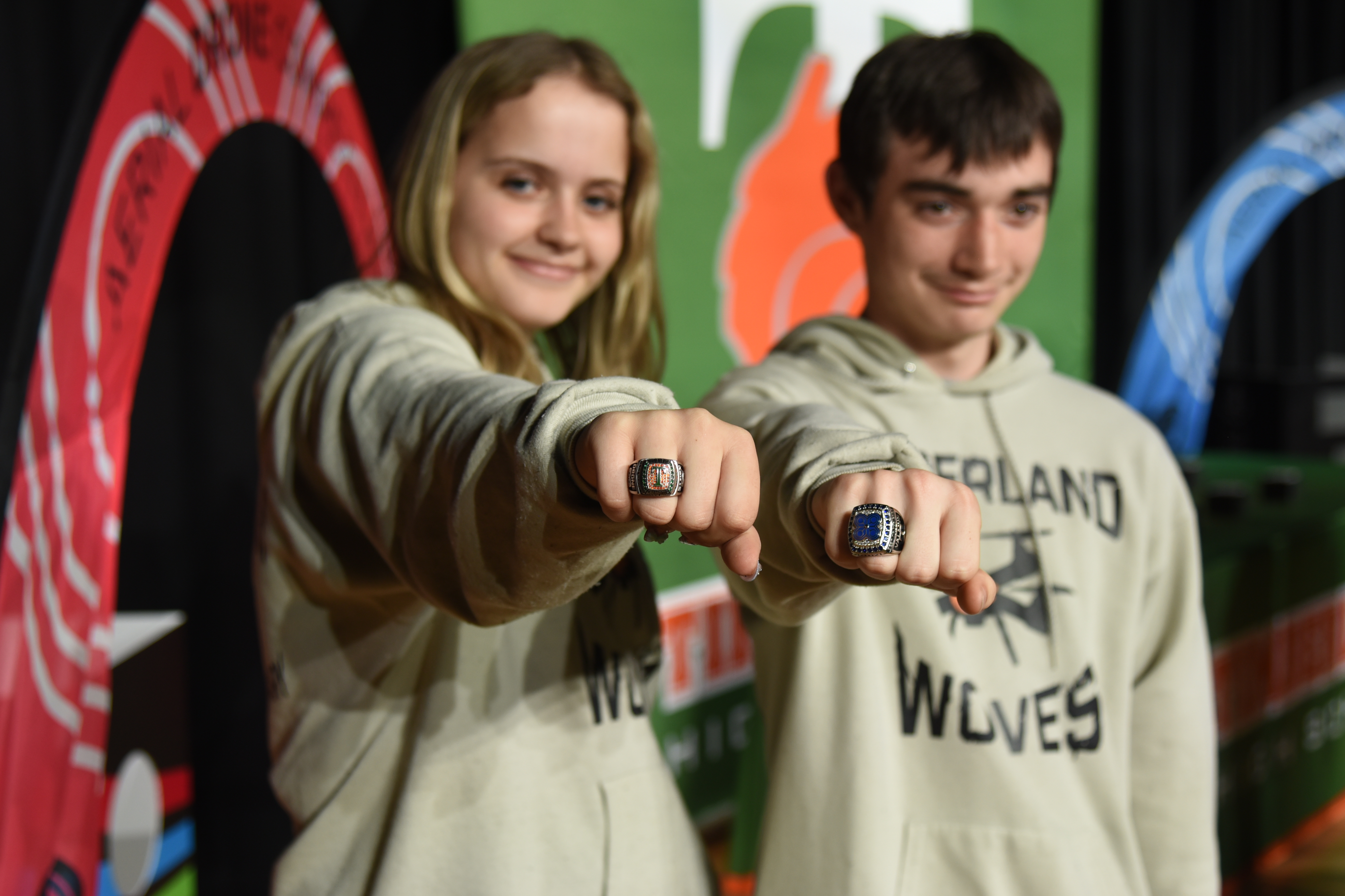 Two individuals in gray hoodies displaying "WOLVES" pose with rings. Behind them, colorful circular designs with letters and numbers are visible.