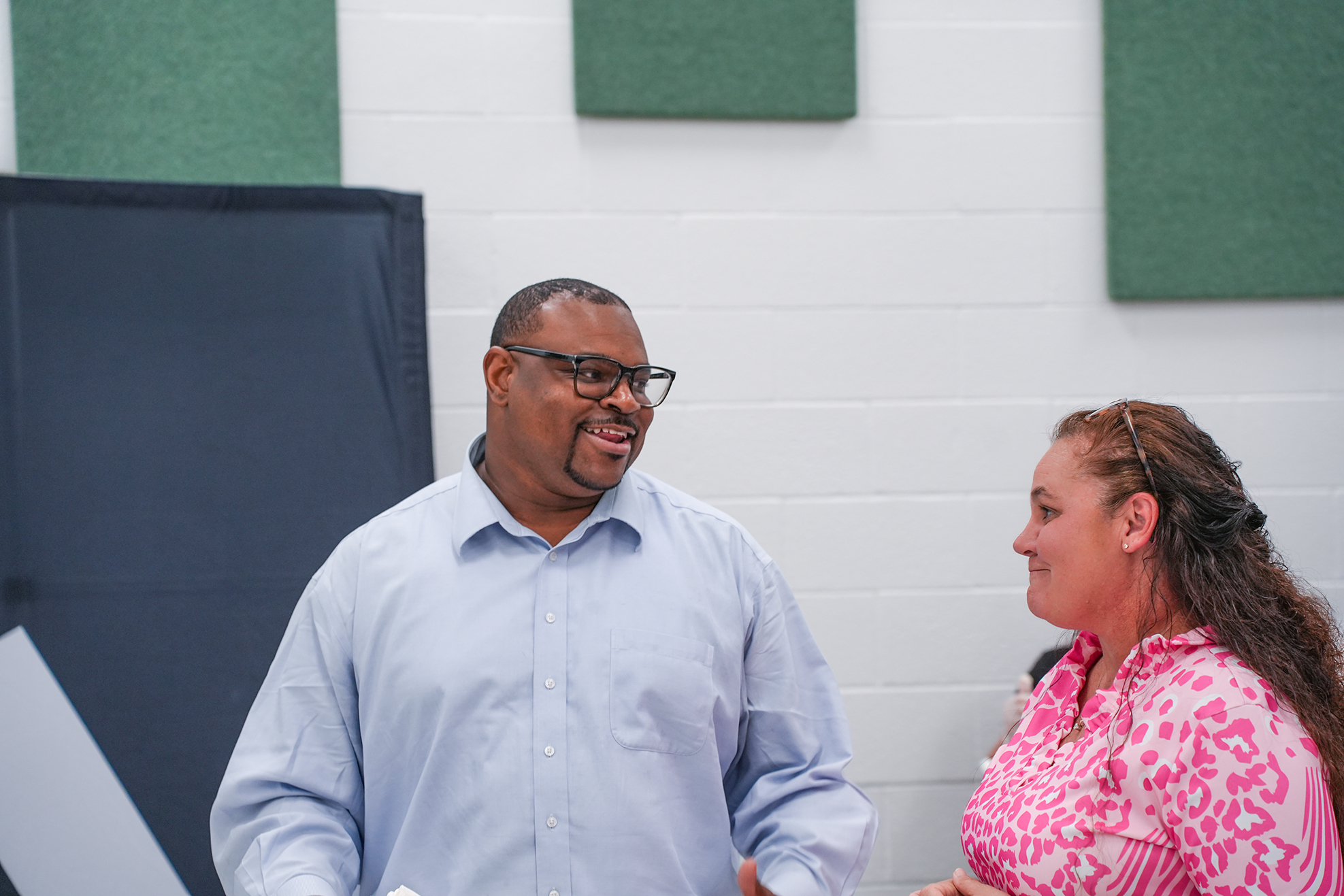 A man and woman in a room with white walls, both smiling. He wears glasses and a blue shirt; she wears a pink blouse.