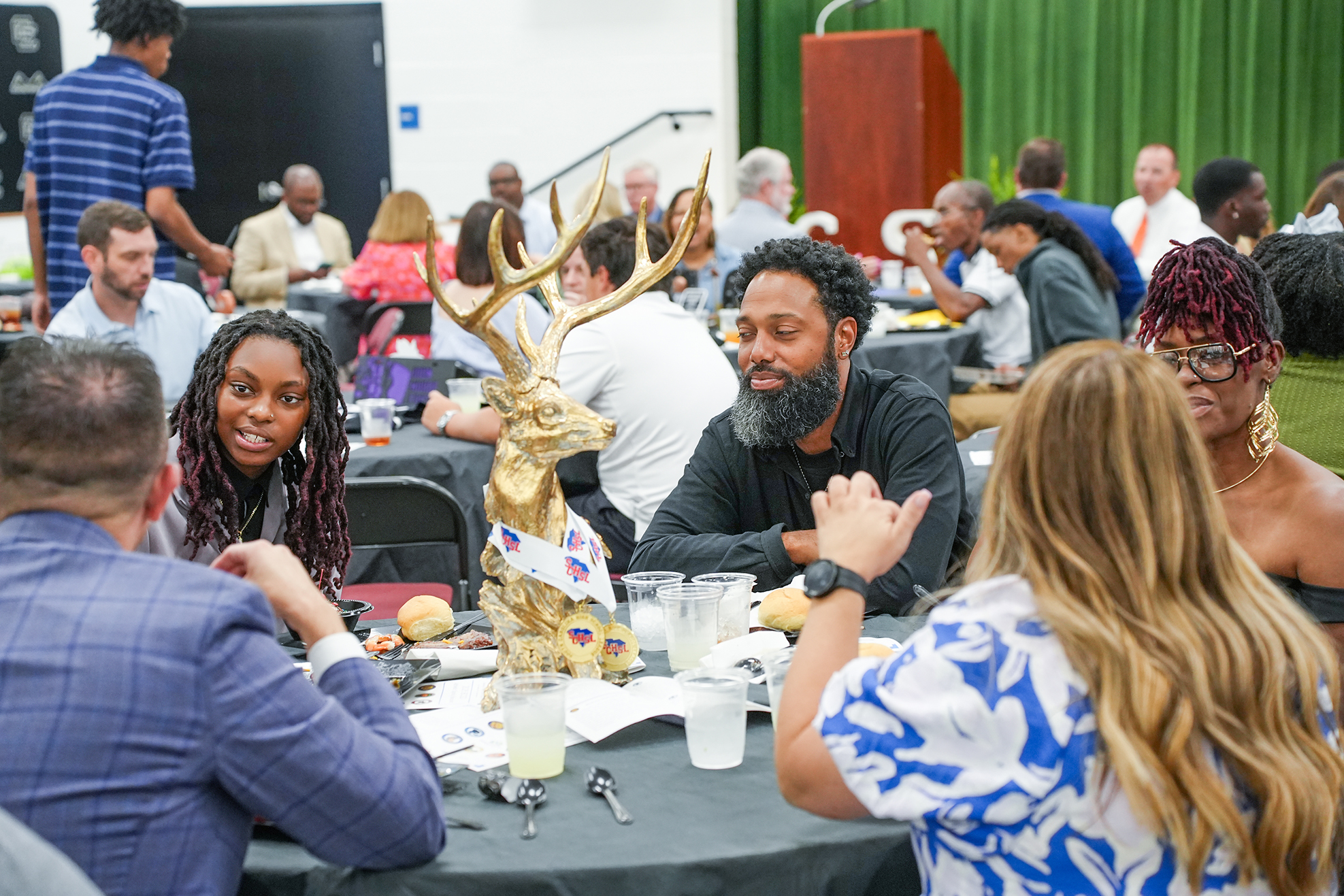 A room with a green curtain background. People sit at tables. A deer statue is on one table.