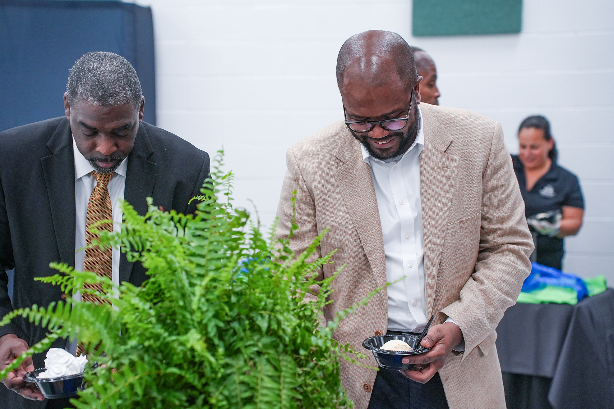 Two men in suits, one holding a bowl of food, standing next to a potted plant.