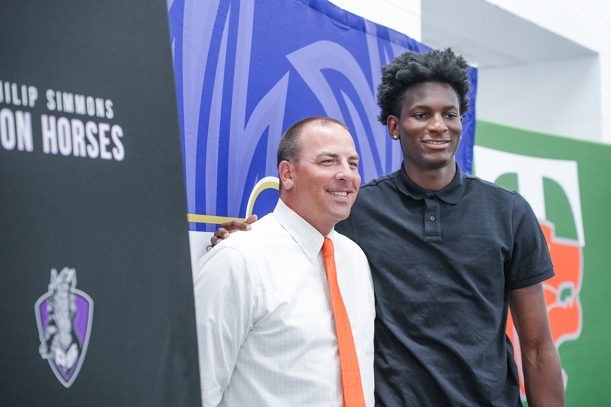 Two men stand smiling, one in a white shirt with an orange tie, the other in a black shirt. Behind them, a backdrop features a logo, text, and a team flag.