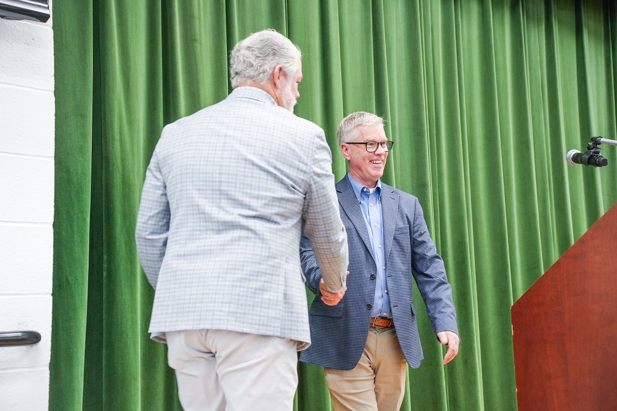 Two men in suits and glasses shake hands near a podium in a room with green curtains.