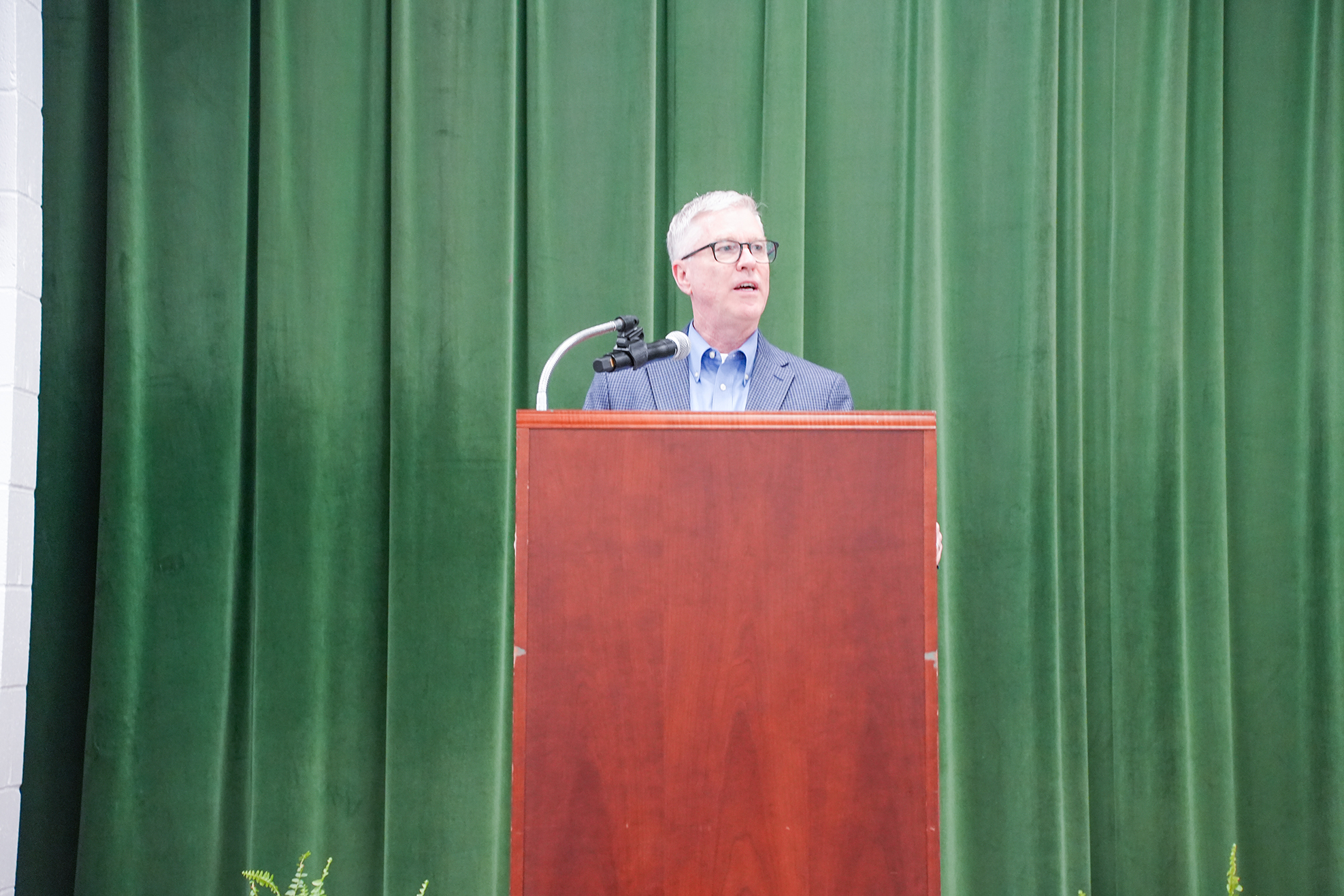 A man in a suit stands at a podium with a microphone, speaking into it. Green curtains are in the background.