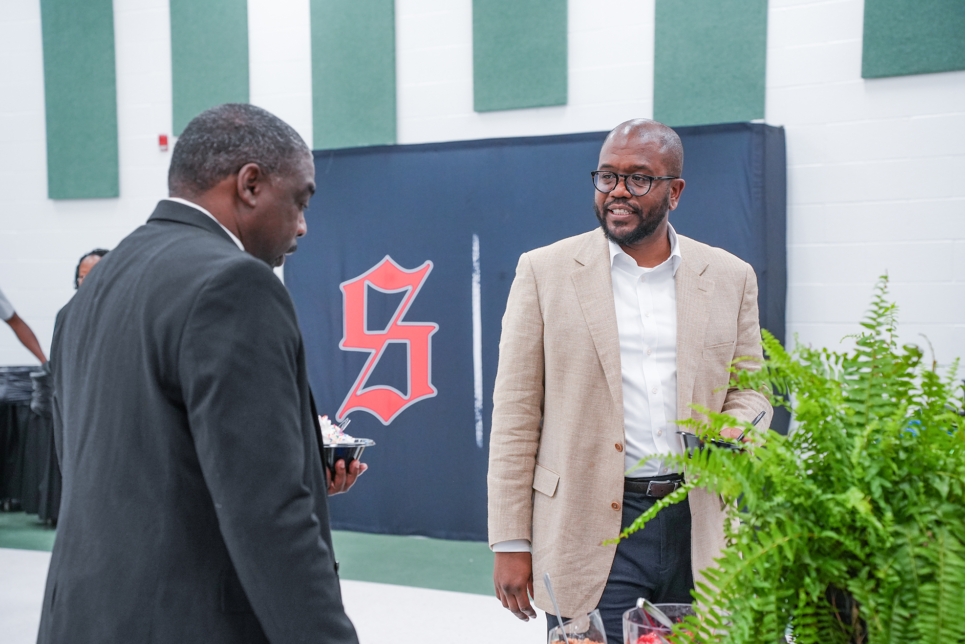 Two men in suits stand next to a banner and plant, facing each other in a room.