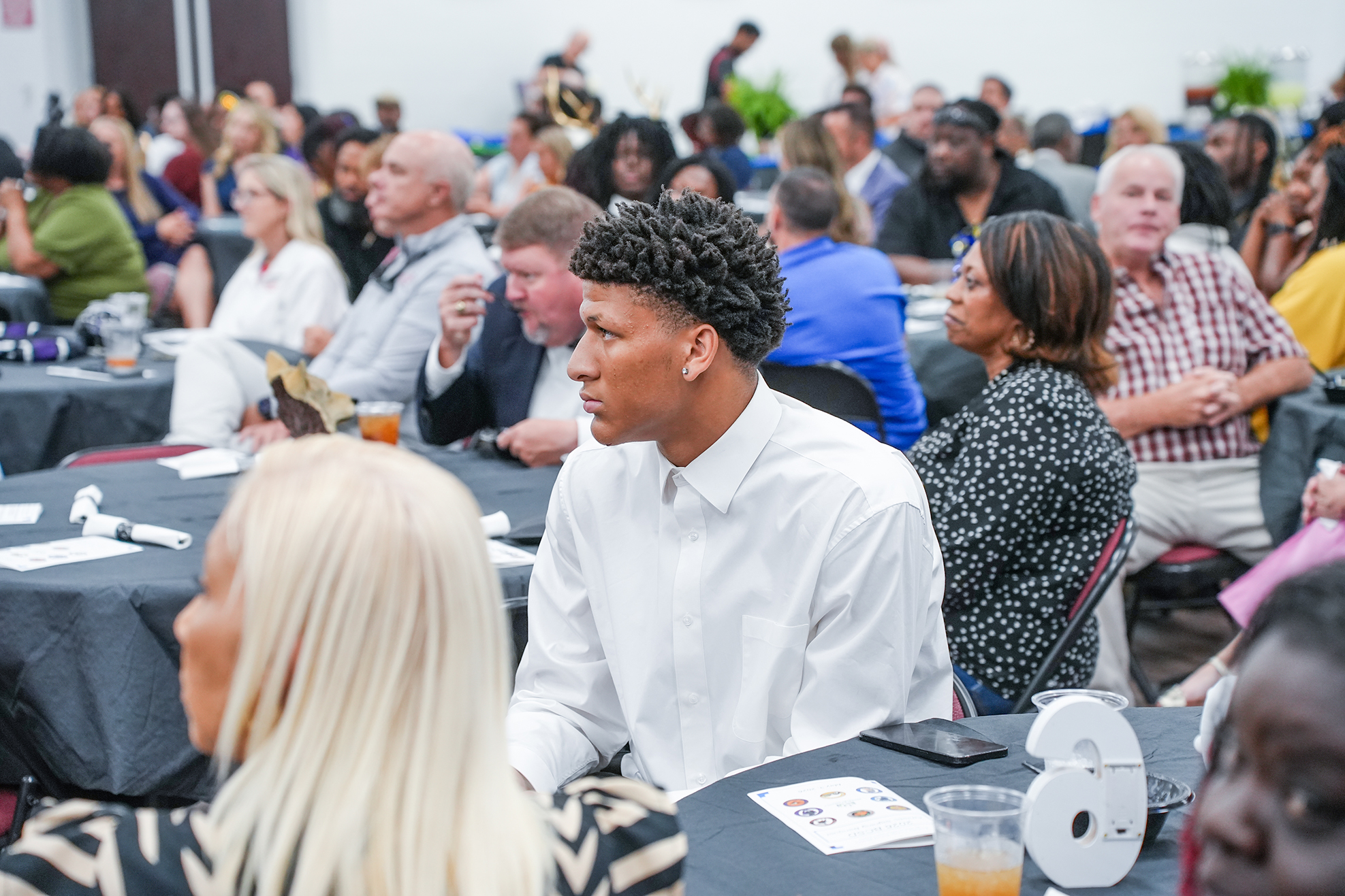 A room filled with people seated at tables. A man in a white shirt speaks to a woman with blonde hair.